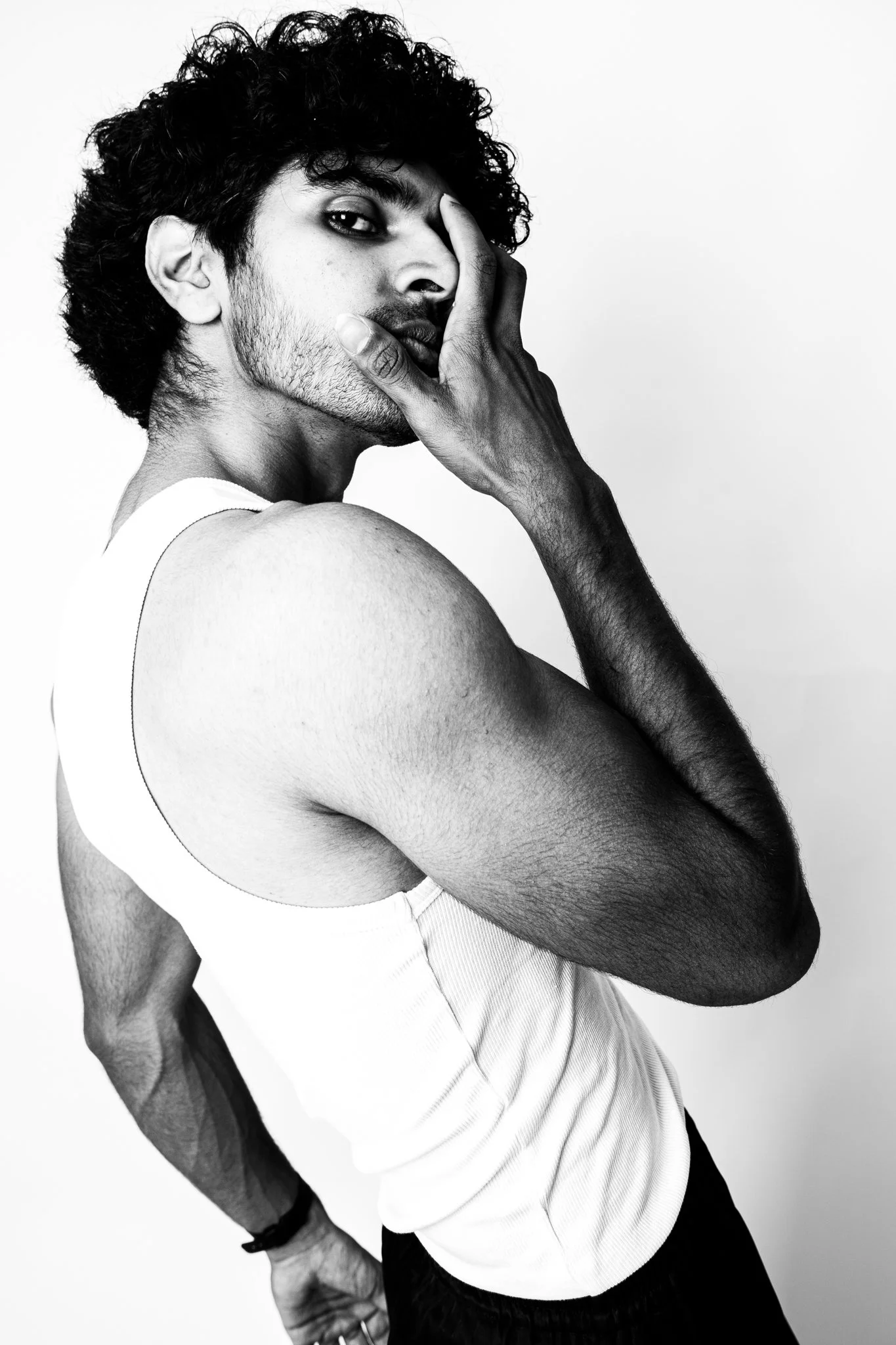 A black and white photo of a young man with dark, curly hair and a beard, wearing a sleeveless white athletic shirt. He poses with his left hand covering part of his face, with a serious expression, against a plain background.