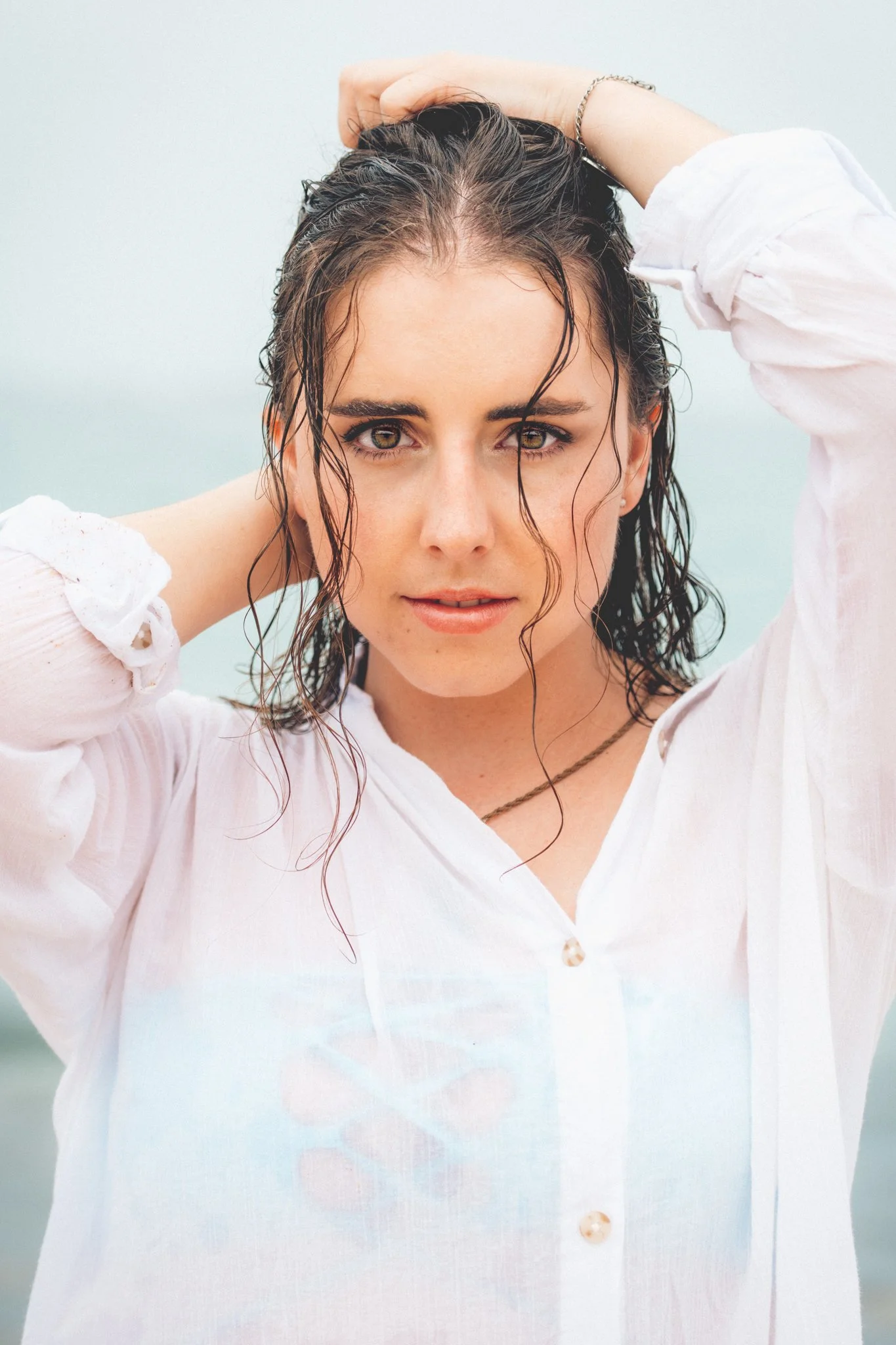 A woman with wet, curly brown hair wearing a white shirt with rolled-up sleeves, standing outdoors near the water, looking directly at the camera with her hands behind her head.