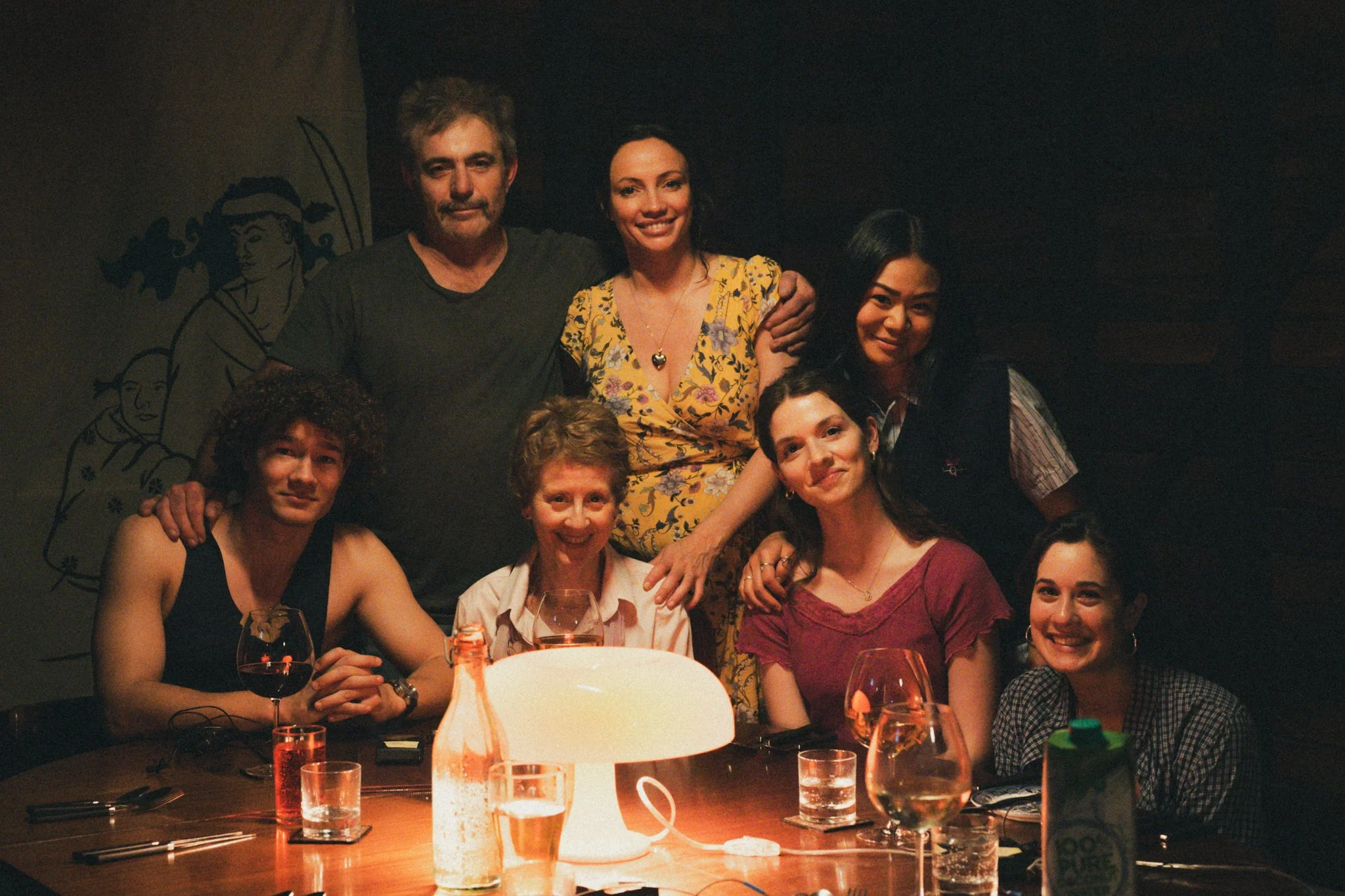 A group of seven people gathered around a dining table with drinks, smiling and posing for the photo in a dimly lit restaurant or bar.