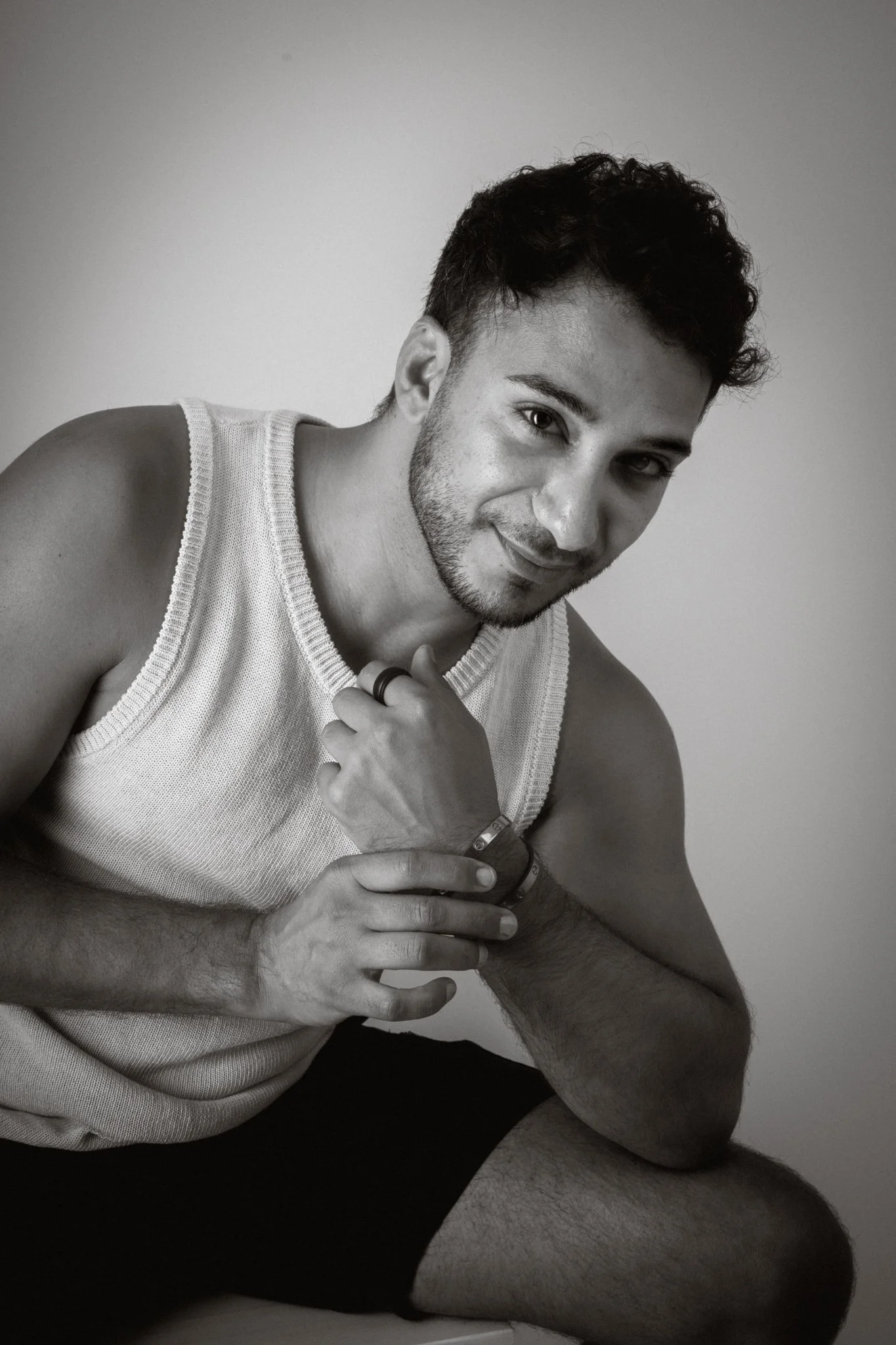 Black and white photograph of a young man with dark, curly hair, a beard, wearing a sleeveless shirt and shorts, sitting against a plain wall, with one knee up, resting his elbow on it, and smiling at the camera.