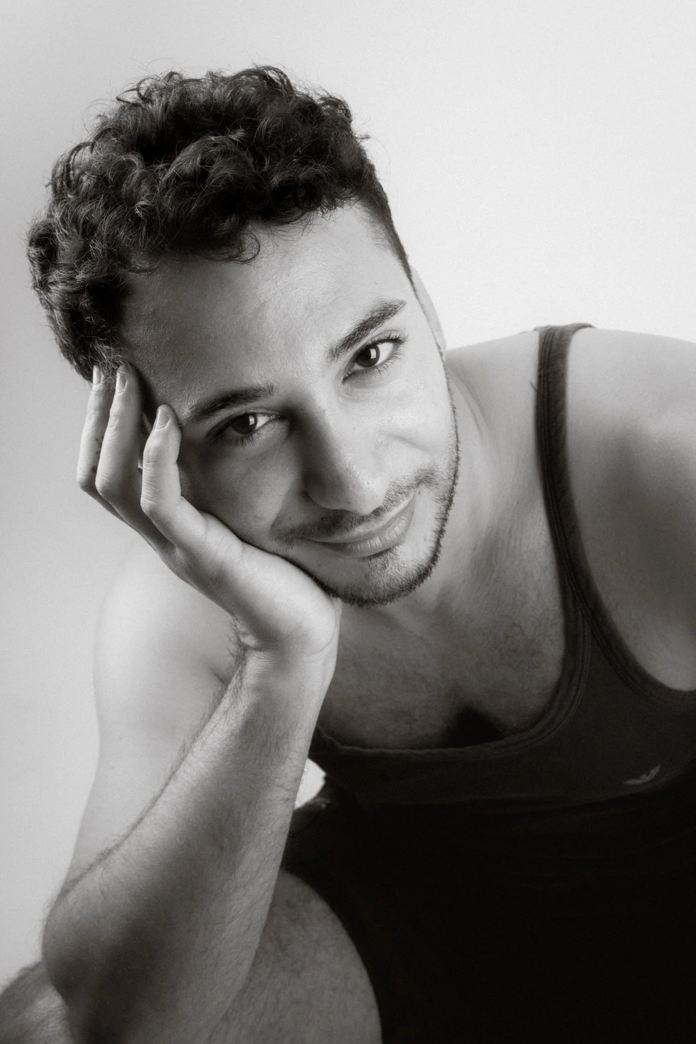 Black and white portrait of a young man with short, curly hair, resting his head on his hand, smiling softly at the camera.