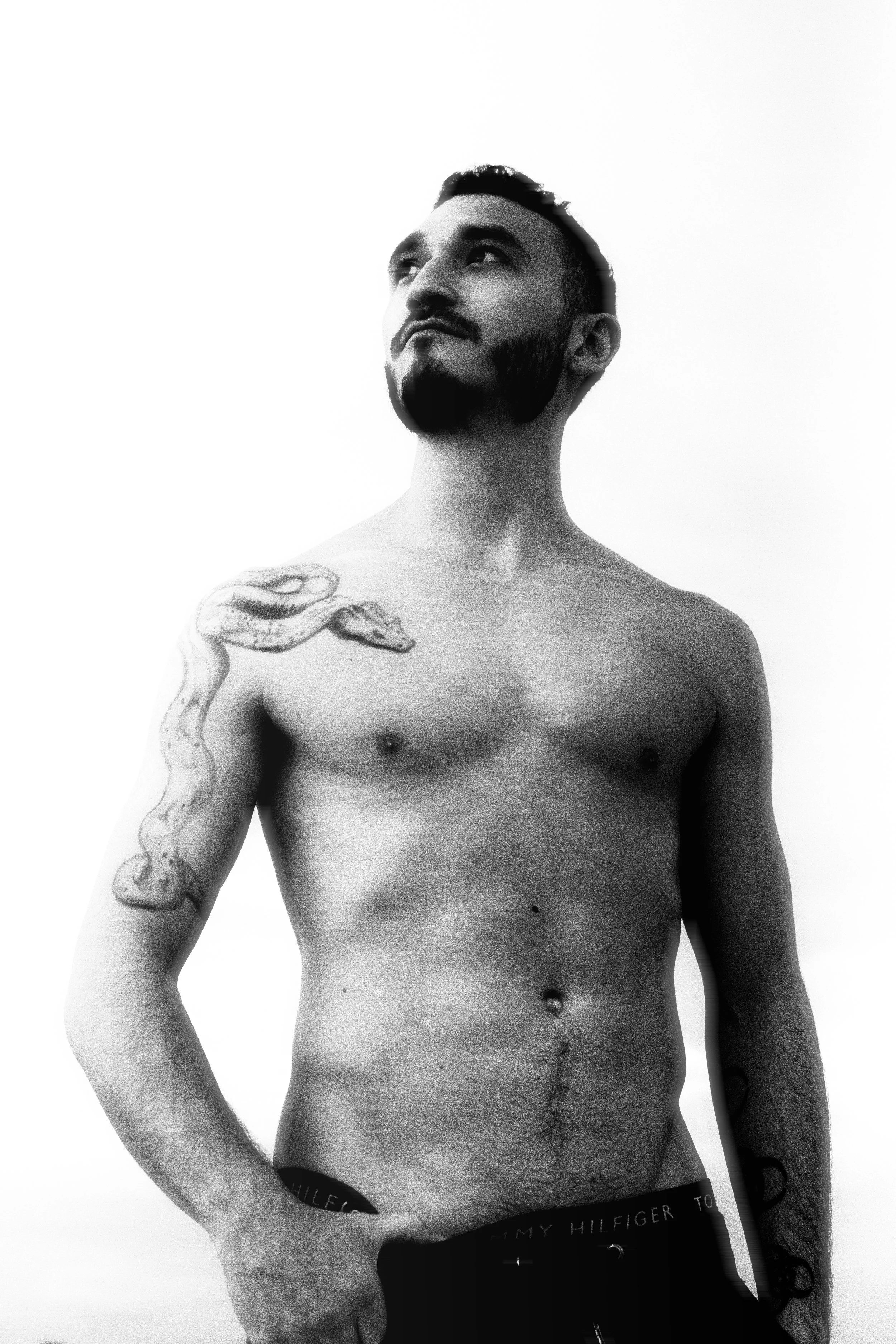 A shirtless man with tattoos, looking upward, with a neutral background.
