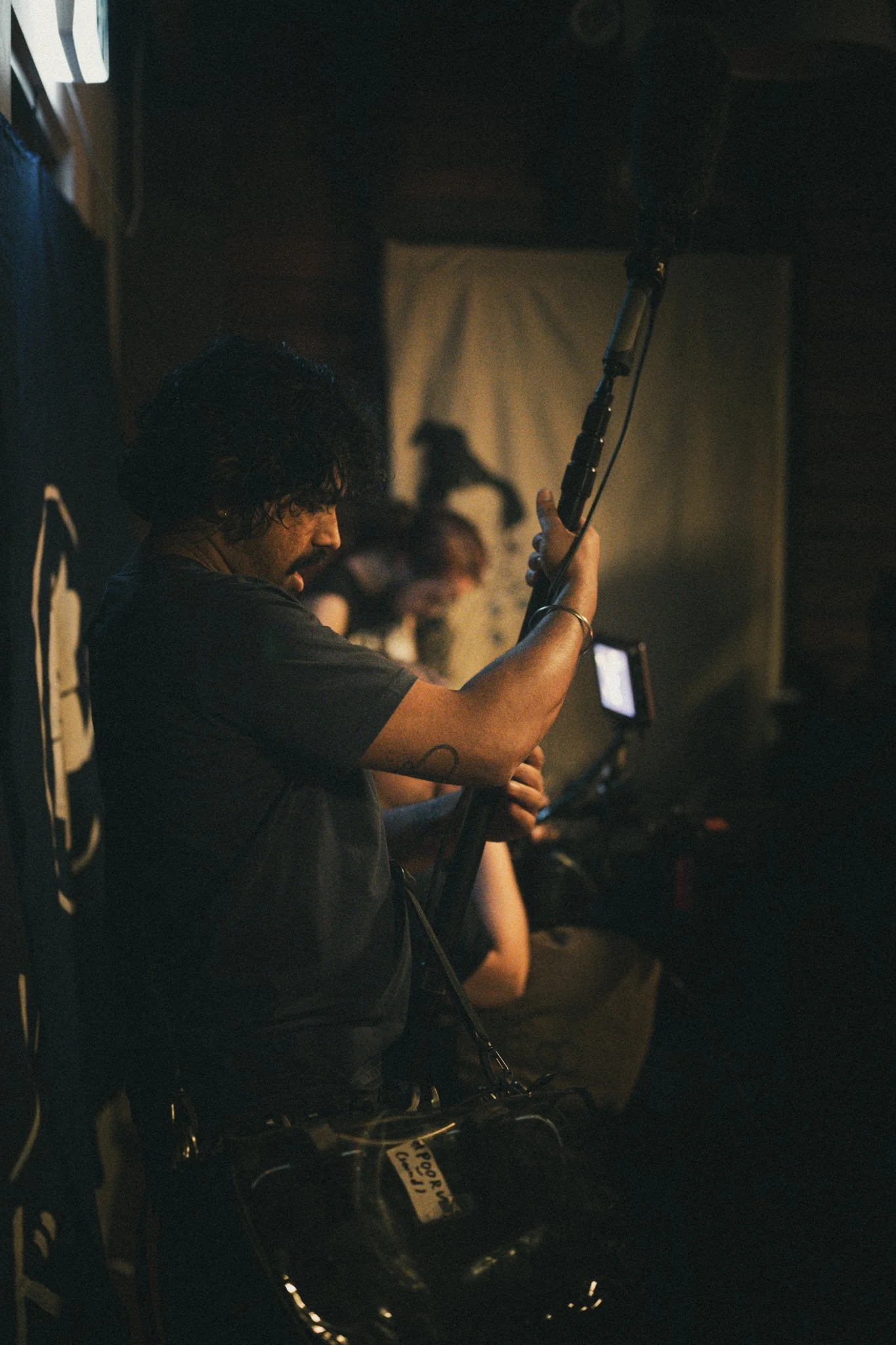 A person holding a boom microphone in a dimly lit indoor setting, with other people in the background engaged in filming or recording activities.