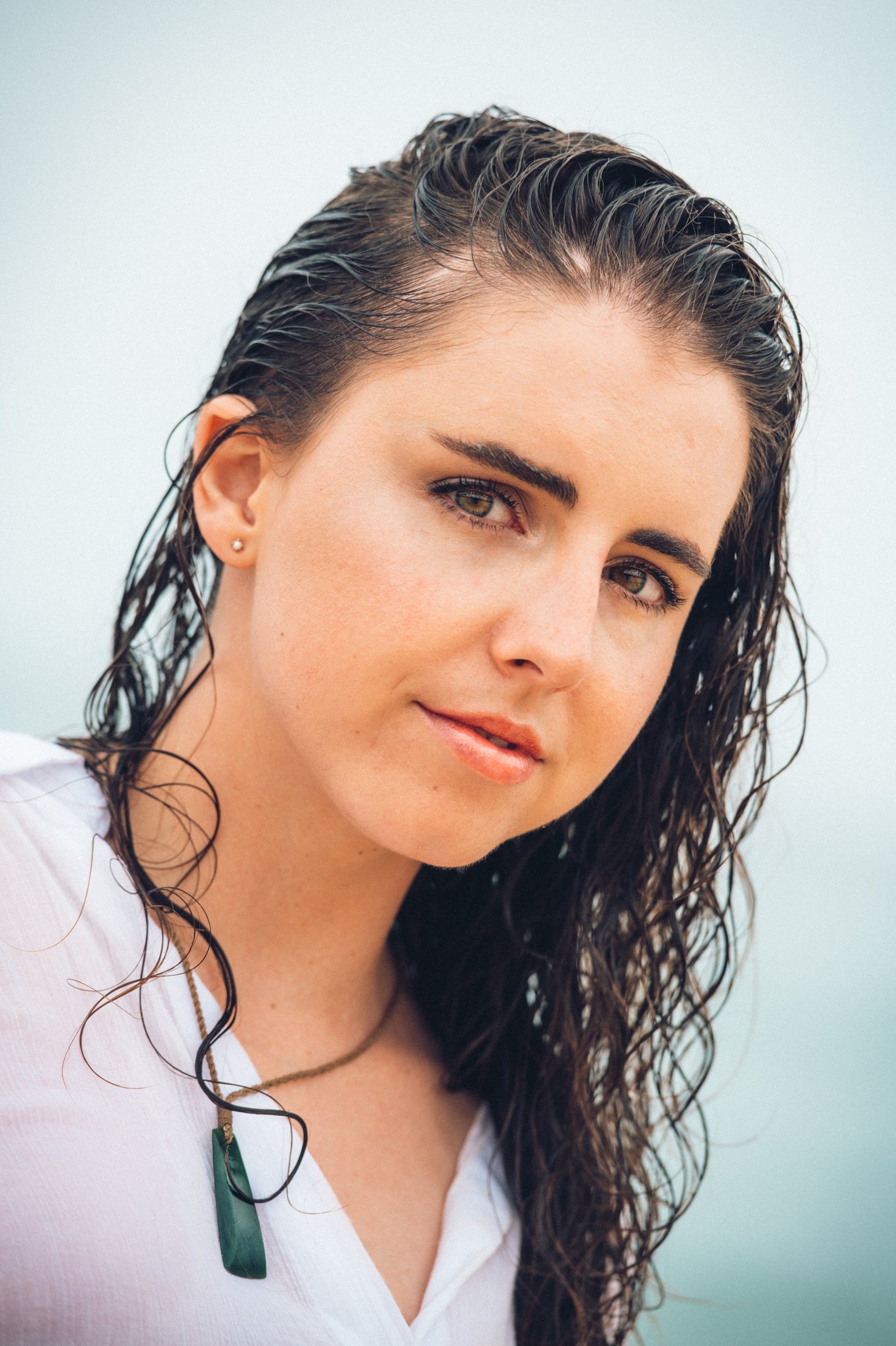 A woman with wet, dark curly hair and light skin, wearing a white top and a green pendant necklace, looking at the camera against a light background.