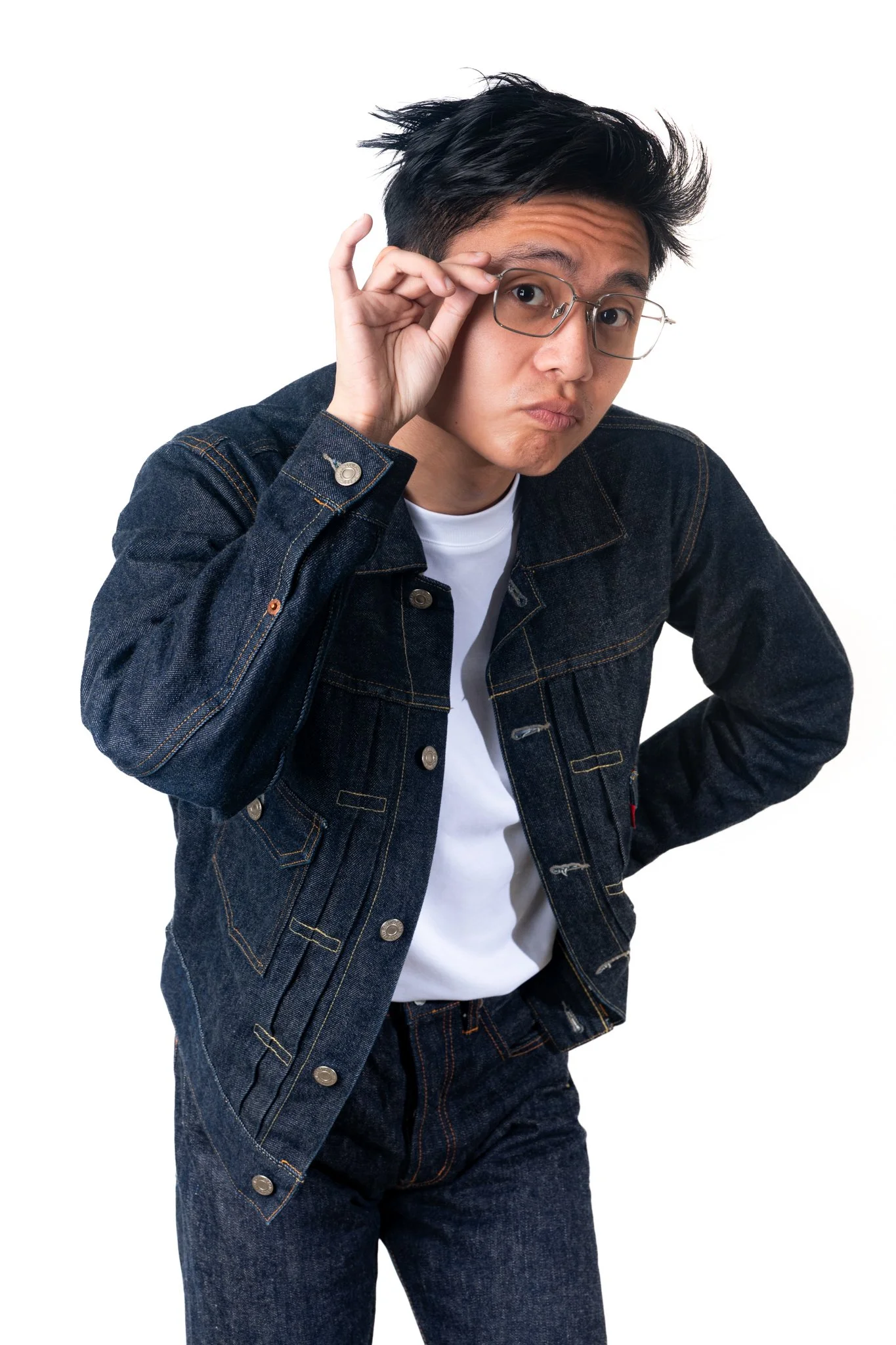 A young man with black hair and glasses, wearing a denim jacket and jeans, looking at the camera with a slightly puzzled expression, adjusting his glasses with his right hand.