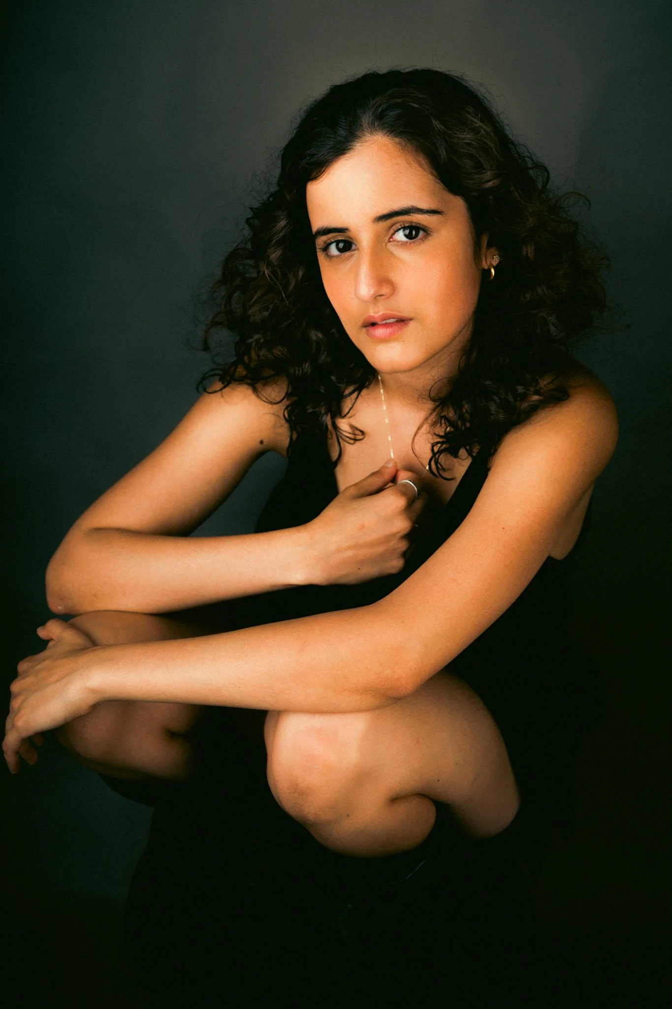 A young woman with curly dark hair wearing a black outfit, sitting against a dark background, looking at the camera.