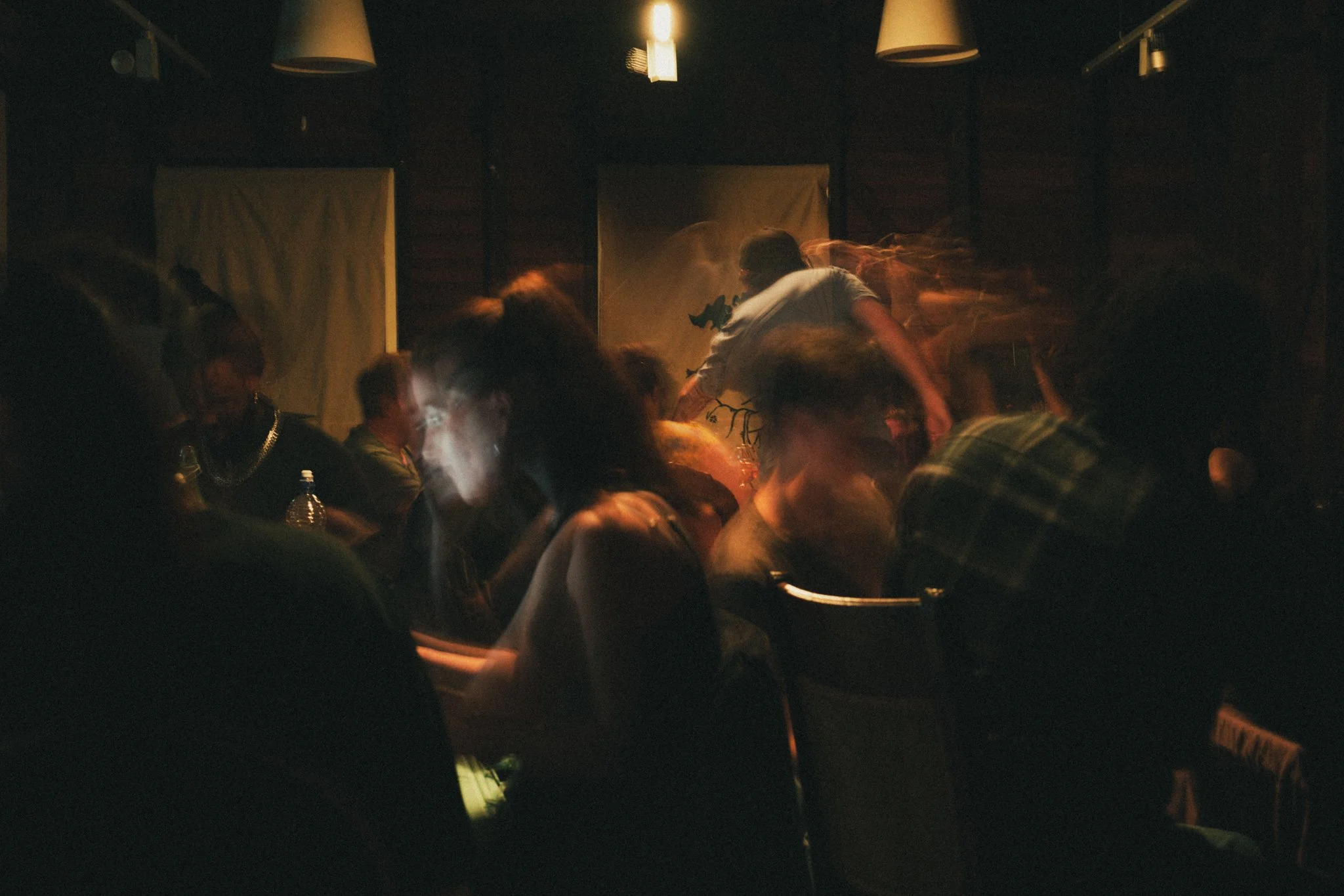Dimly lit indoor setting with people seated around tables, one person is standing and reaching across the table, with a blurry motion effect indicating movement, and a warm light fixture hanging from the ceiling.