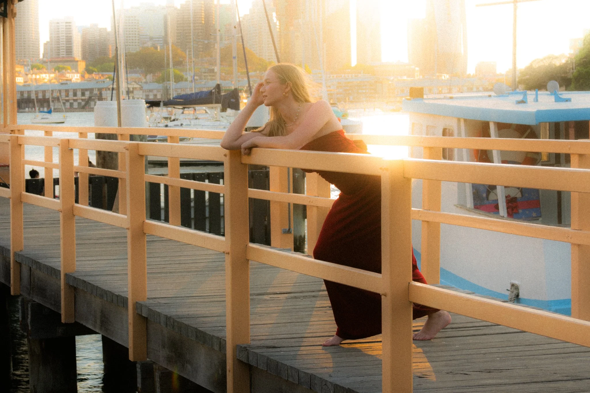 A woman in a red dress leaning on a wooden railing on a dock, with a marina and city skyline in the background during sunset.