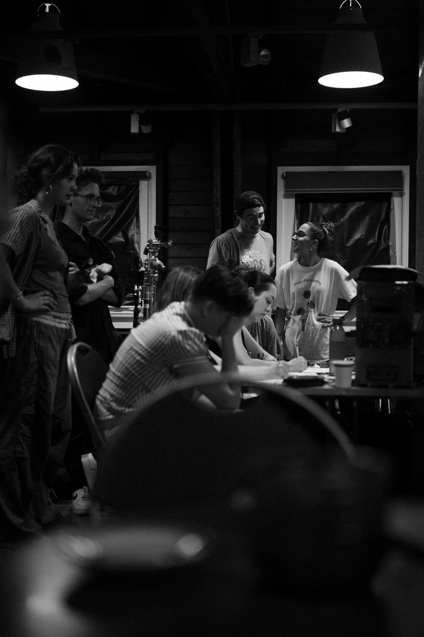 Group of people working on a film set in a dimly lit room with wooden walls and hanging lights.