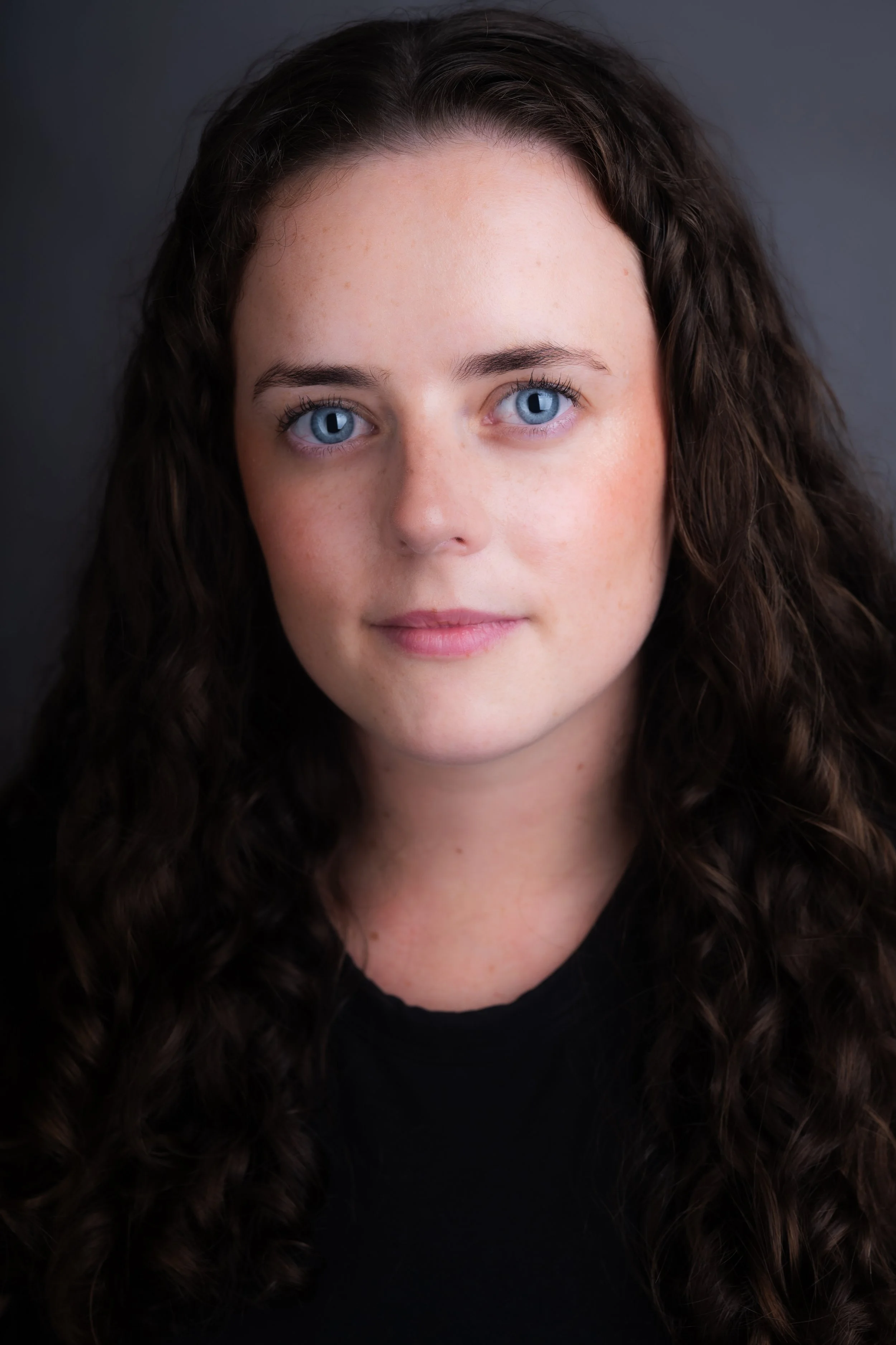 Close-up portrait of a woman with blue eyes and long, curly brown hair, wearing a black top, against a dark background.