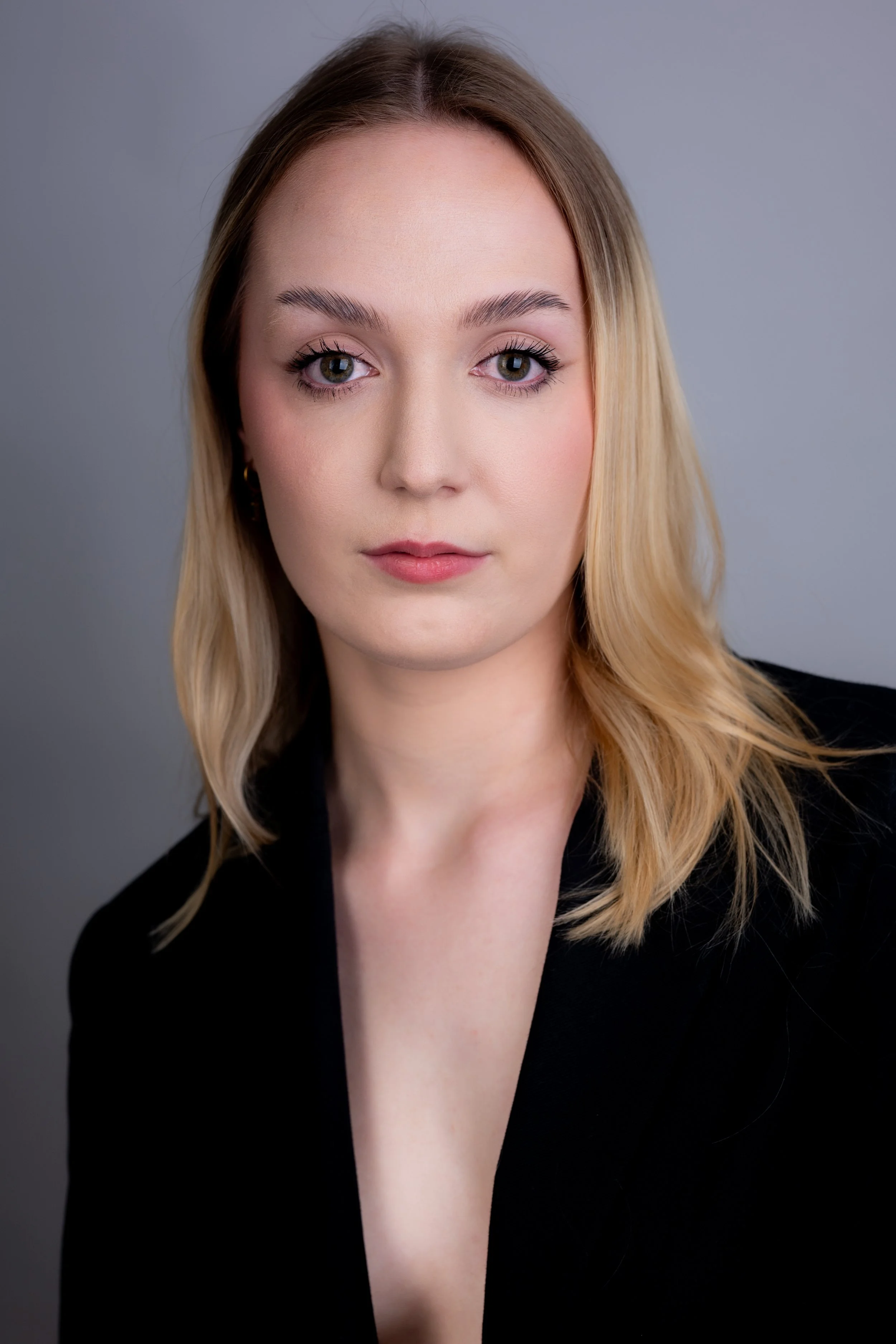 Close-up portrait of a young woman with blonde hair and blue eyes, wearing a black blazer, against a neutral gray background.