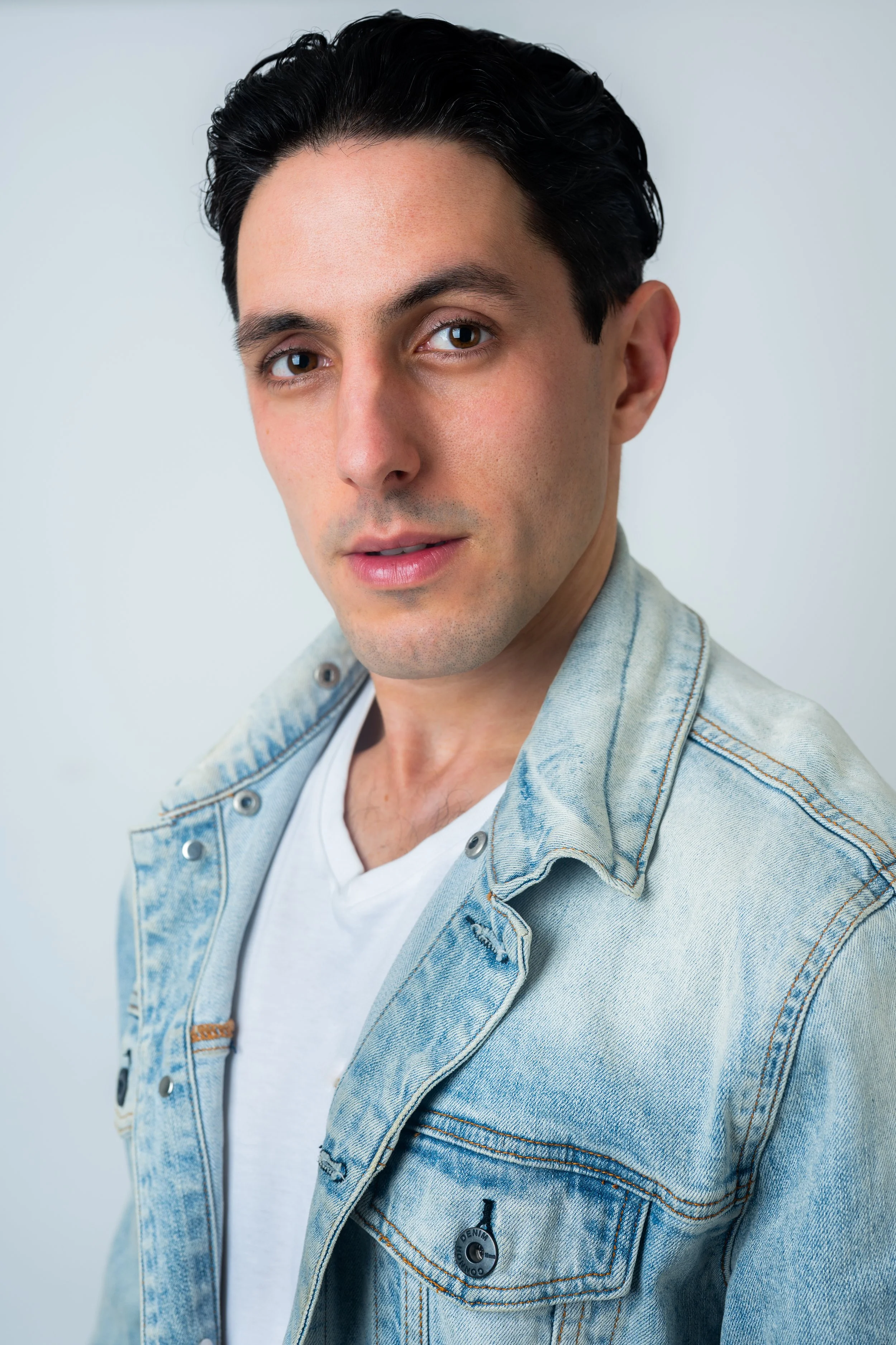 A young man with dark hair and fair skin, wearing a light denim jacket over a white T-shirt, posing against a plain light background.