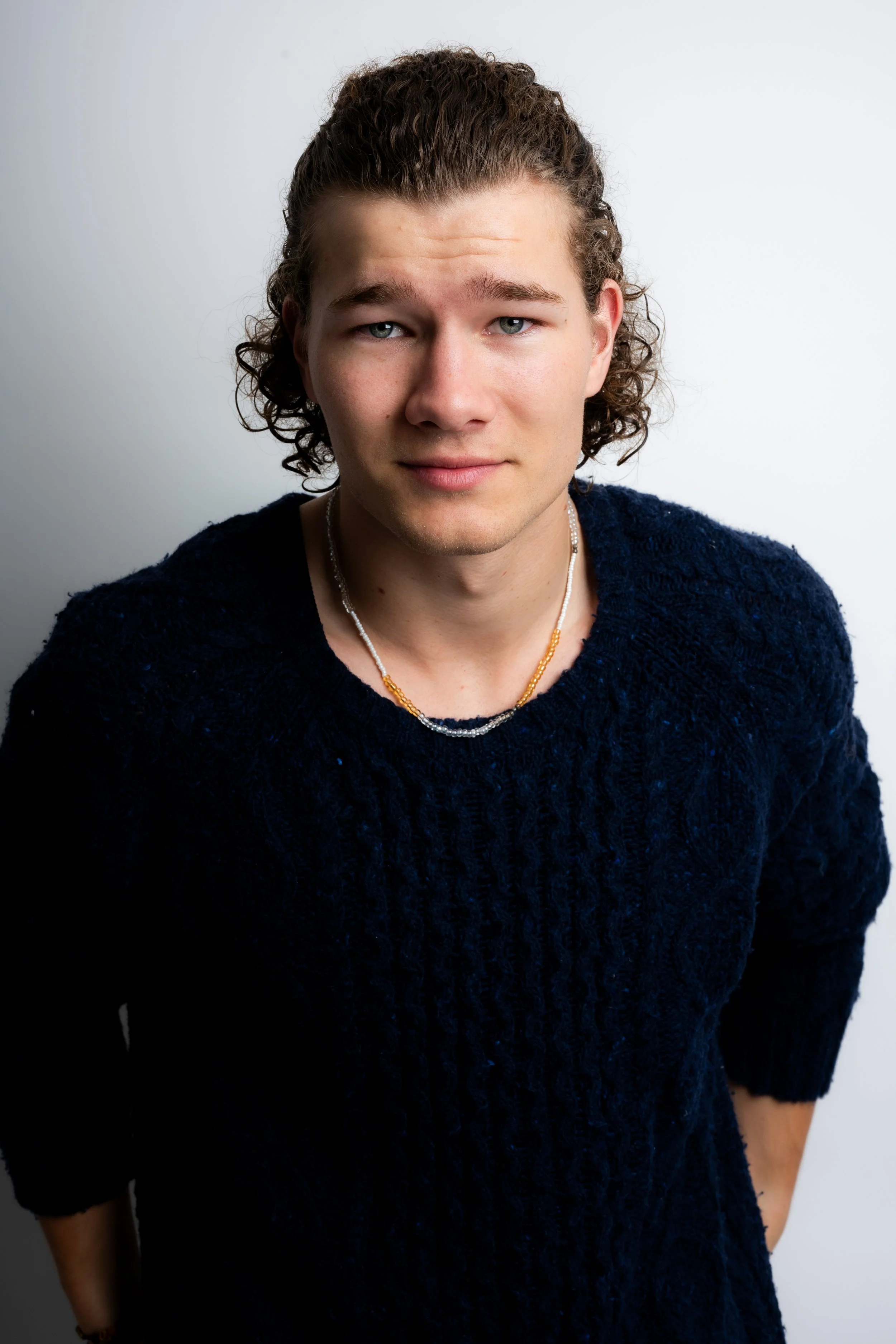 Portrait of a young man with curly hair wearing a navy blue sweater and a beaded necklace, looking into the camera against a plain white background.