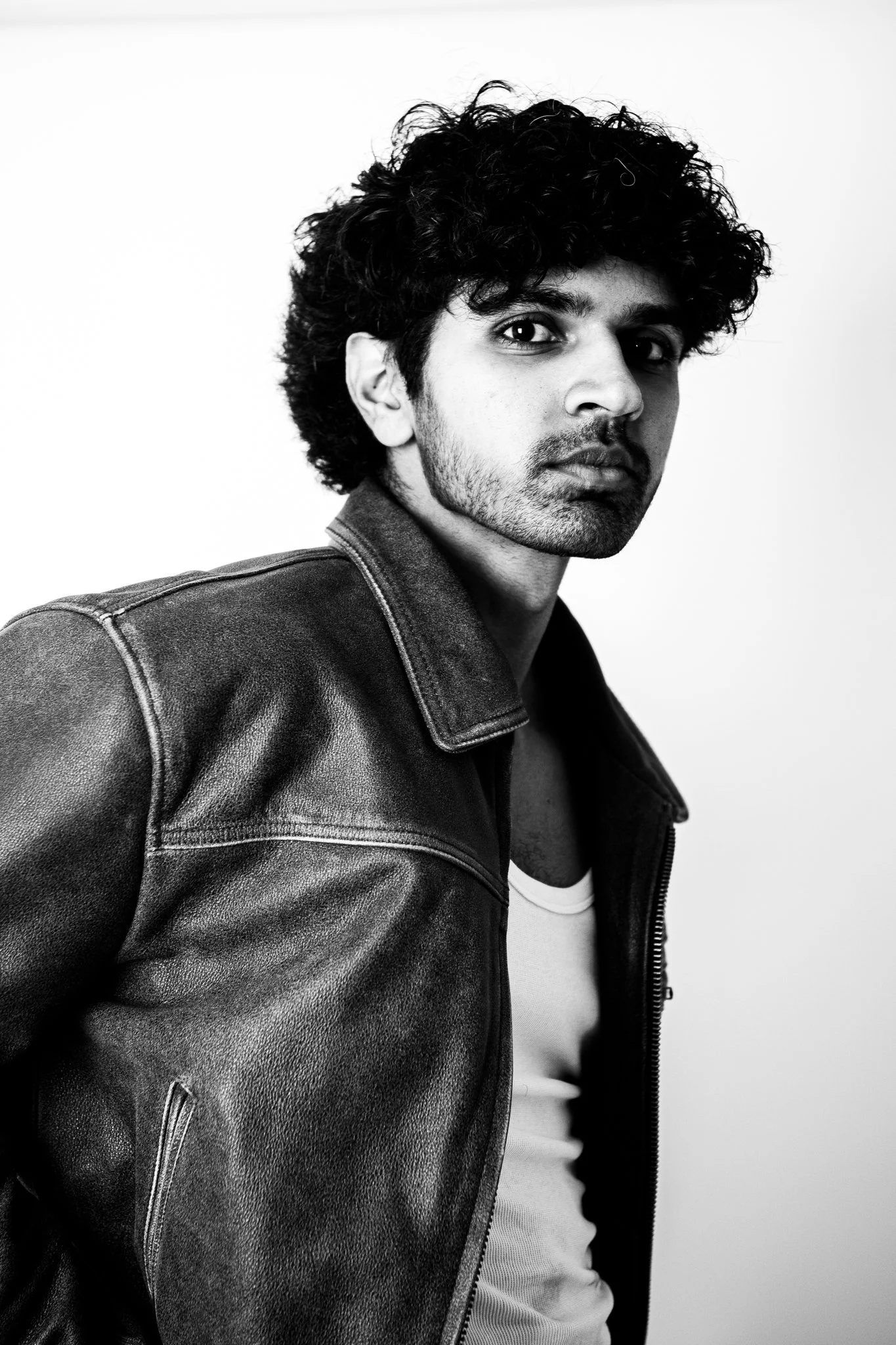 Black and white portrait of a young man with curly hair, wearing a leather jacket and a white shirt, against a plain background.