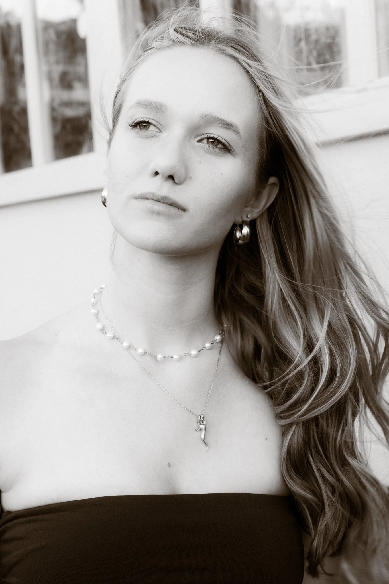 Black and white portrait of a young woman with long, wavy hair, wearing jewelry including earrings and necklaces, looking slightly upward and to the side, with a serious expression.