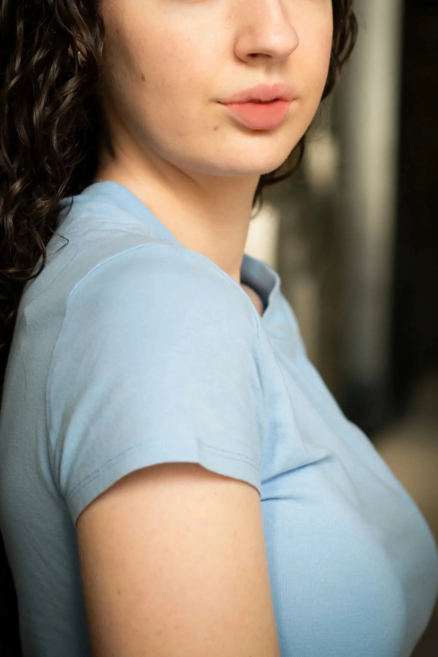 Close-up of a woman's face and shoulder, wearing a light blue shirt, with curly brown hair, against a blurred background.