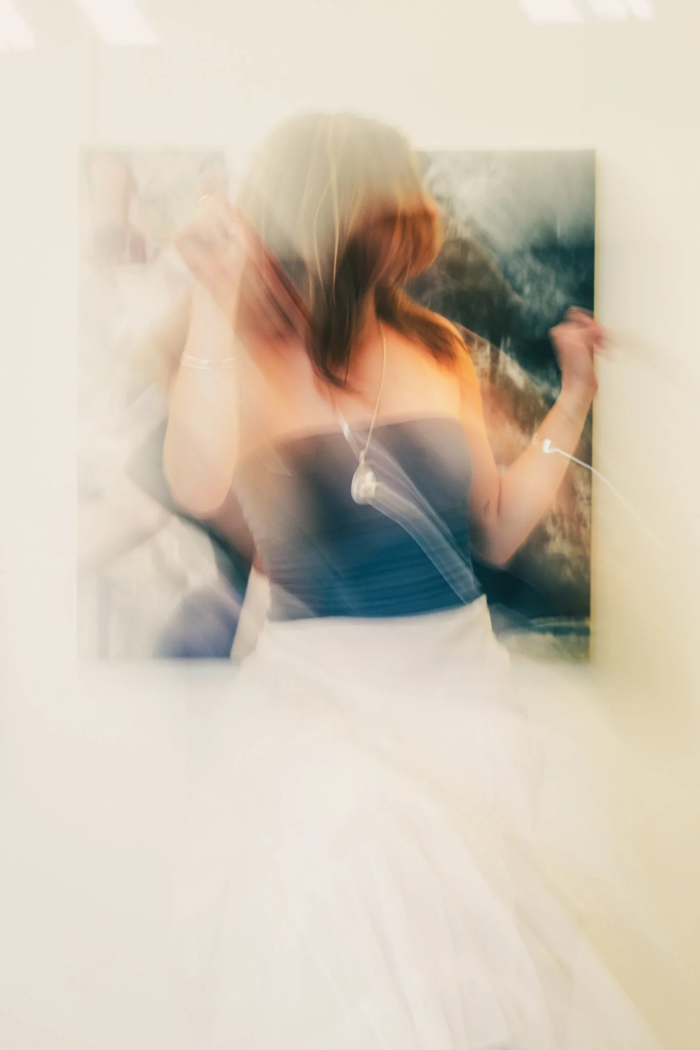 Blurry photograph of a woman with brown hair wearing a black strapless top and white skirt, holding a phone in one hand and a cigarette in the other, standing in front of a painting.