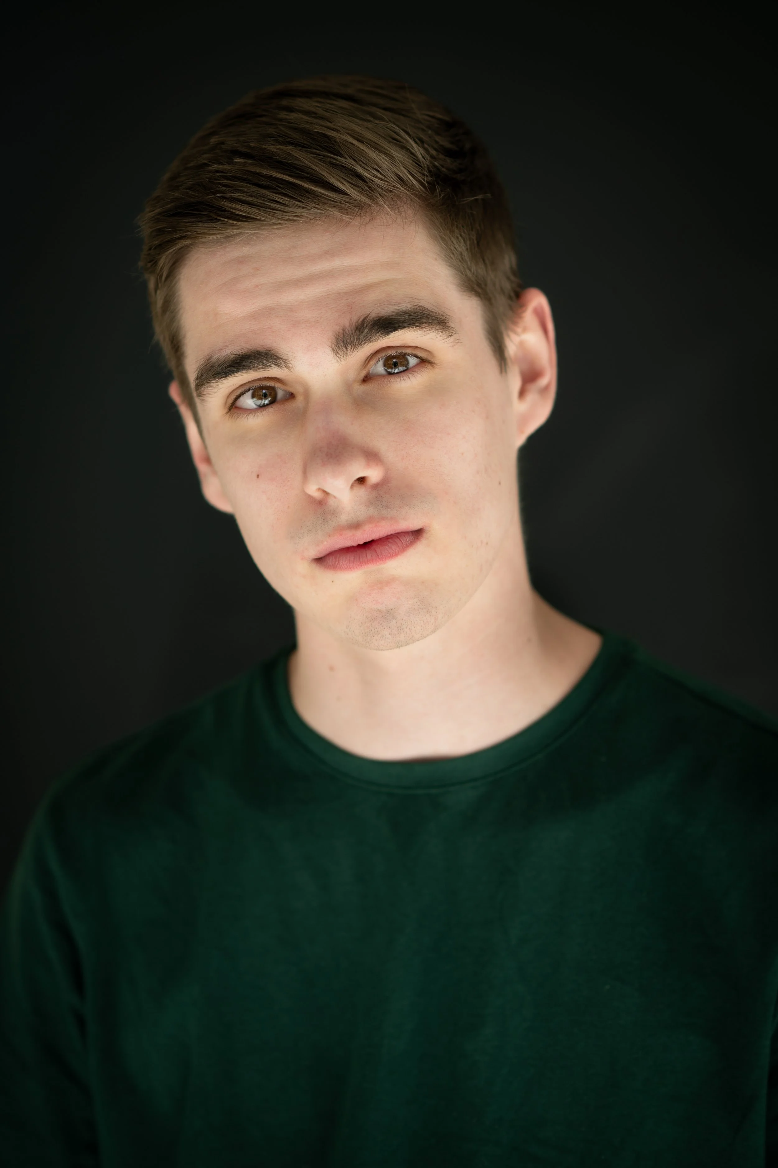 Close-up portrait of a young man with light skin and brown hair, wearing a dark green shirt, against a black background.