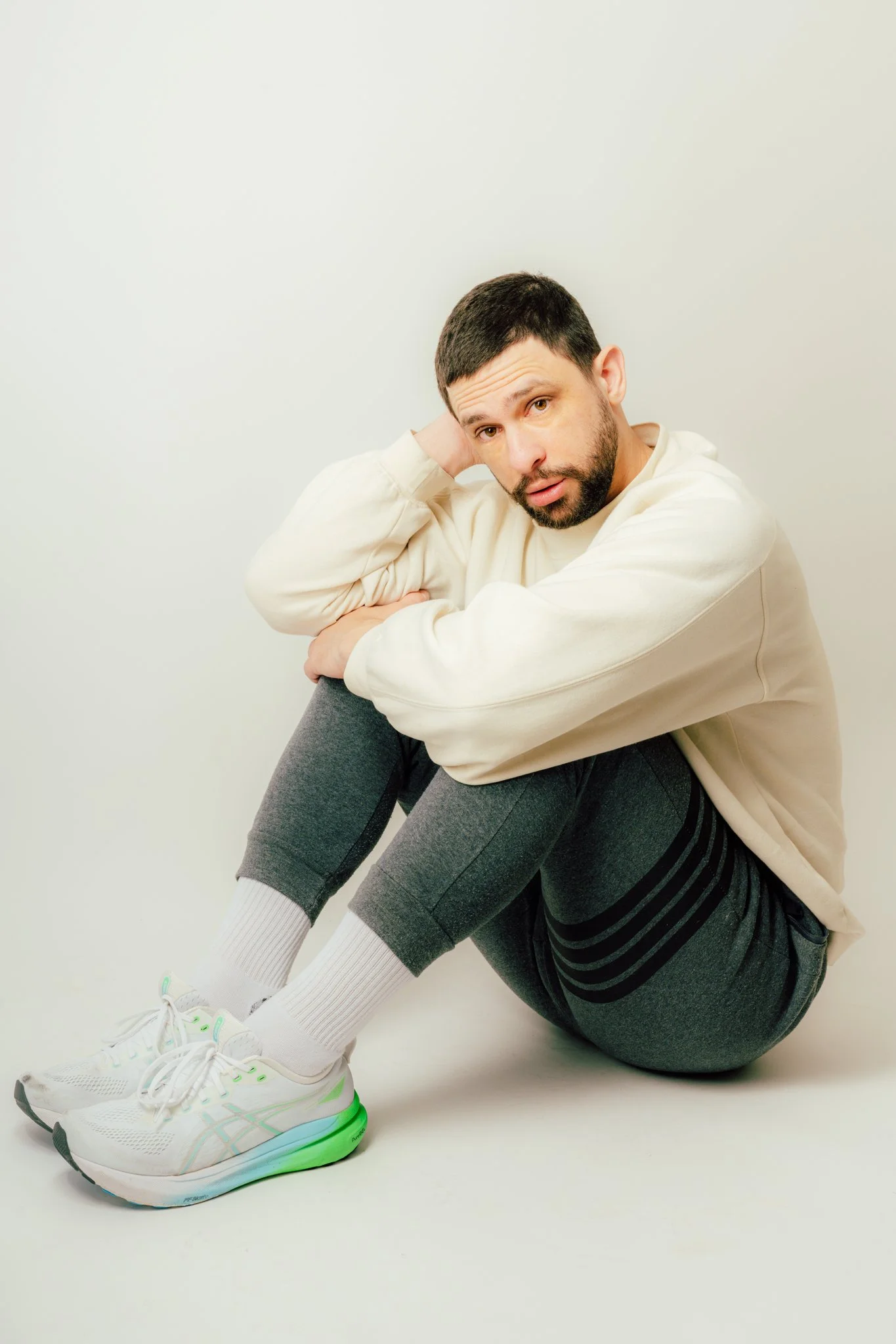 Man with dark hair and beard sitting on the floor, leaning his head on his hand, wearing a beige sweatshirt, gray sweatpants, white socks, and white sneakers with green accents against a plain white background.