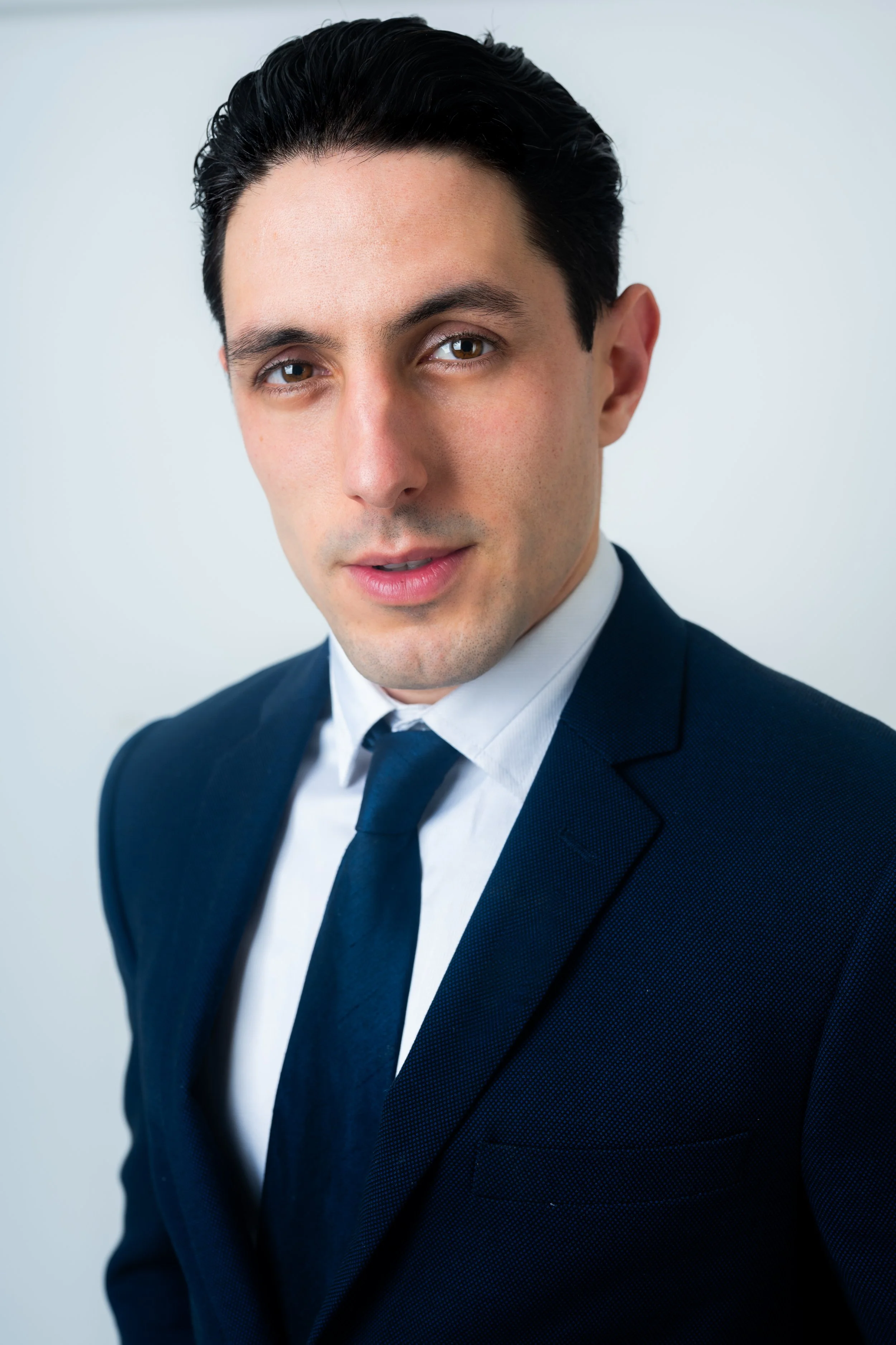 Headshot of a young man in a dark suit, white shirt, and dark tie, with black hair styled back, against a light background.