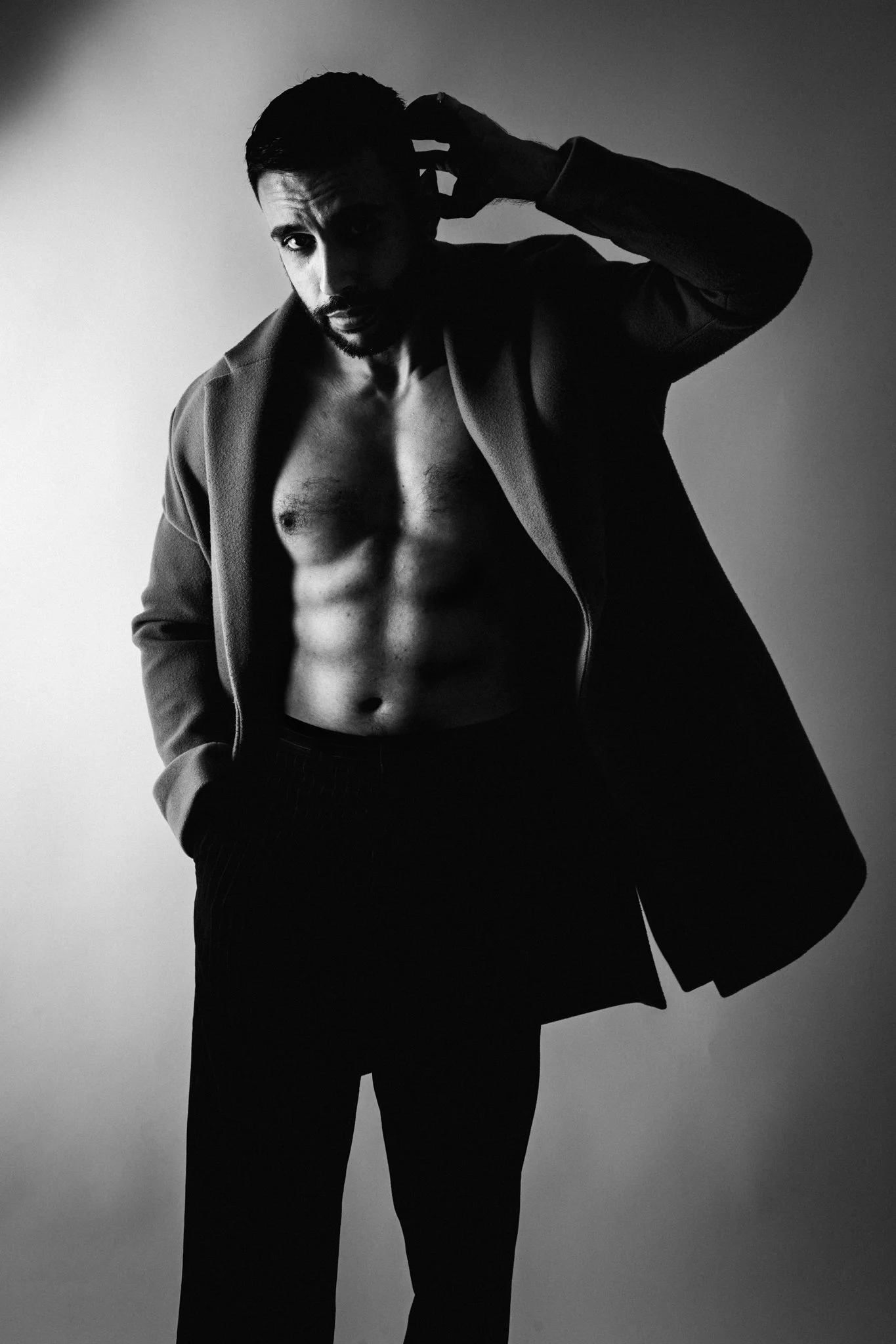 A black and white photo of a shirtless man with a blazer, posing with one hand behind his head and looking at the camera.