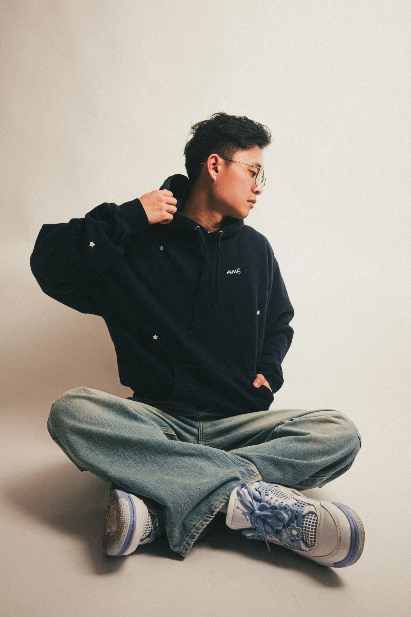 A person sitting cross-legged on the floor, wearing jeans, sneakers, and a dark hoodie, looking to the side against a plain, light background.