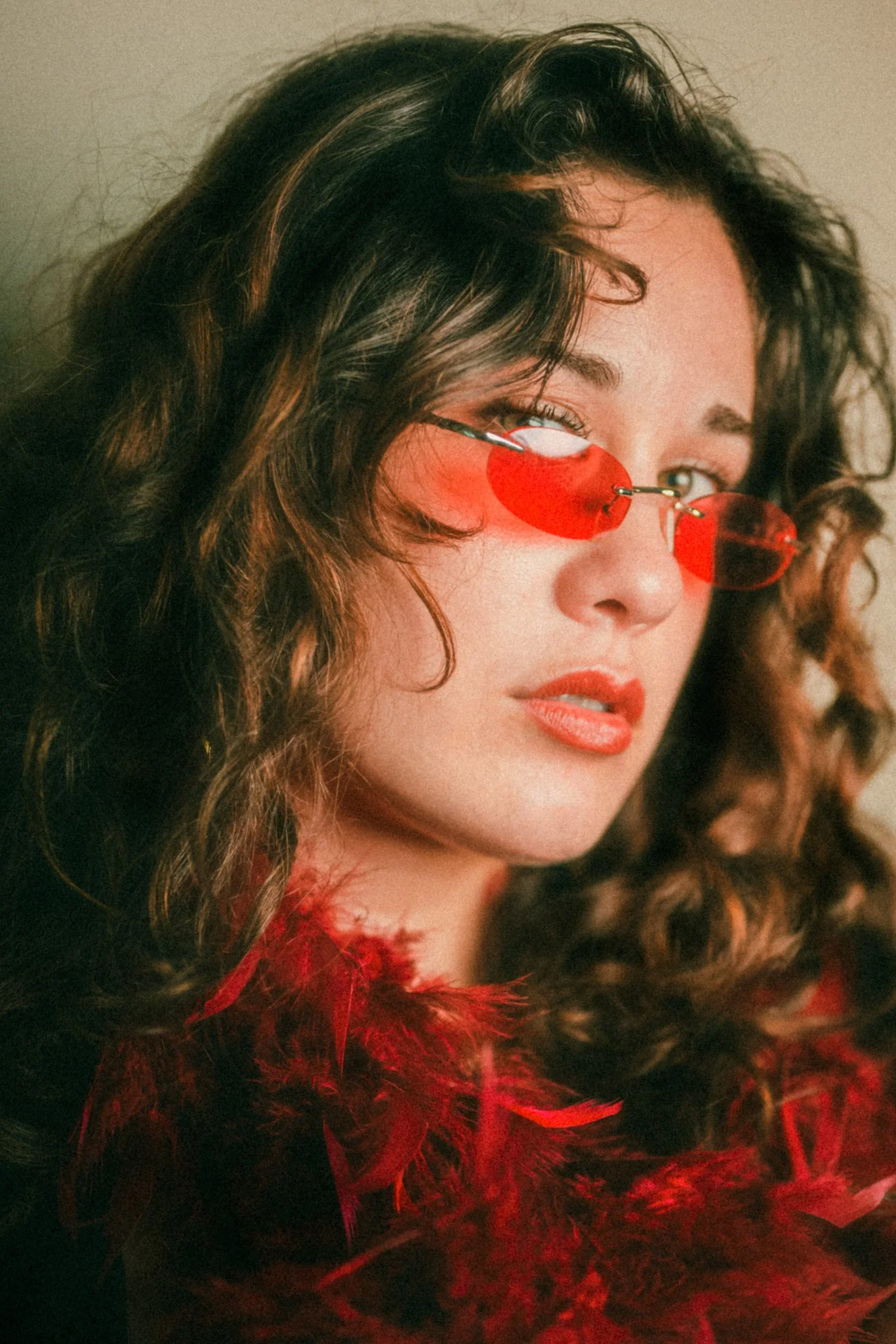 Close-up of a woman with curly dark hair, wearing small red sunglasses, red lipstick, and surrounded by red feathers.