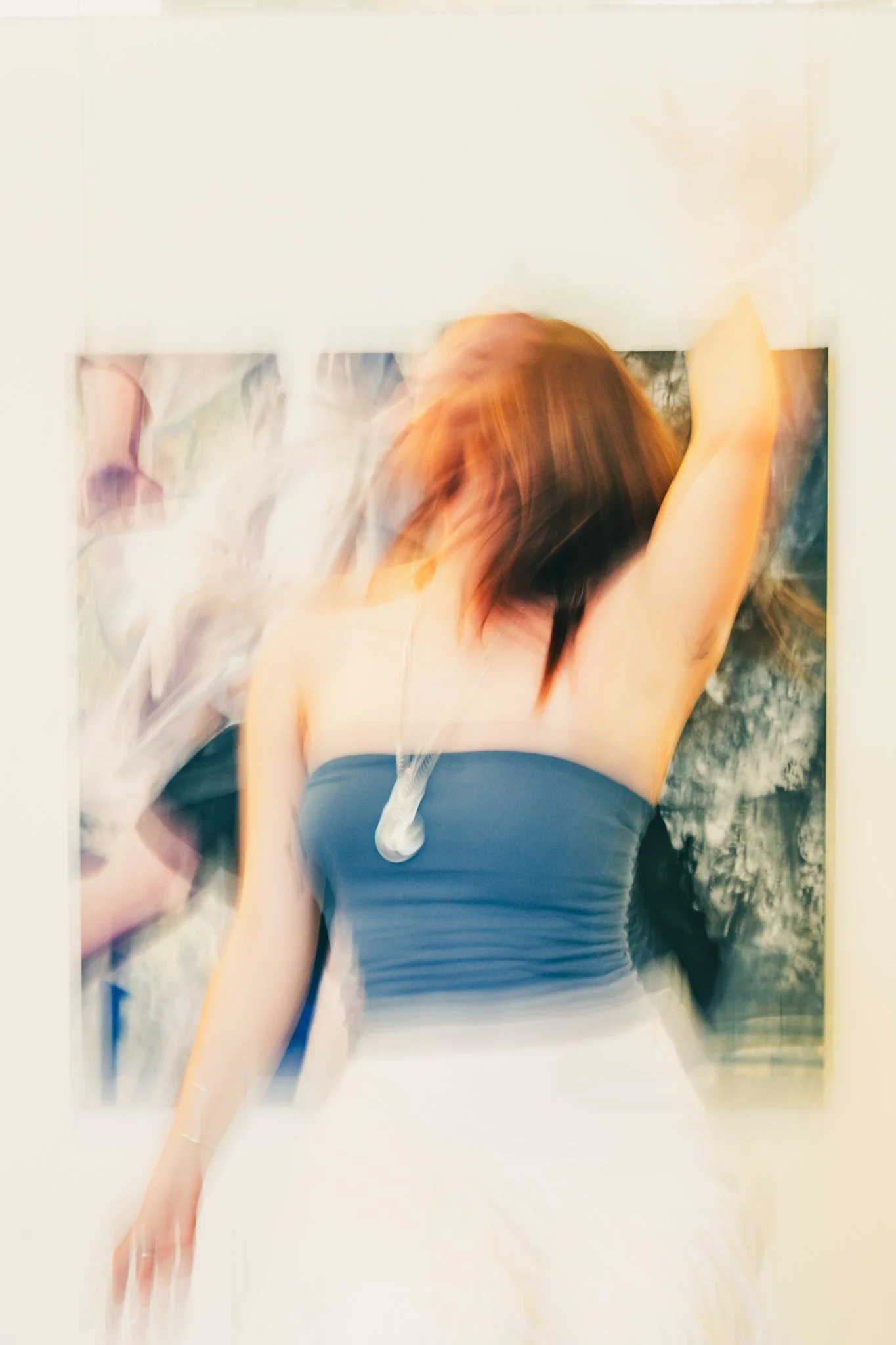 A woman with reddish-brown hair in a strapless blue top, raising her arm, standing in front of abstract wall art, with motion blur.