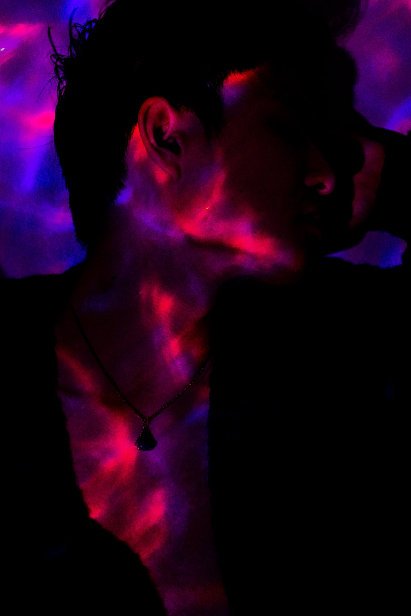 A person with short hair and a necklace in a dark setting with colorful, swirling light patterns on their face and neck.