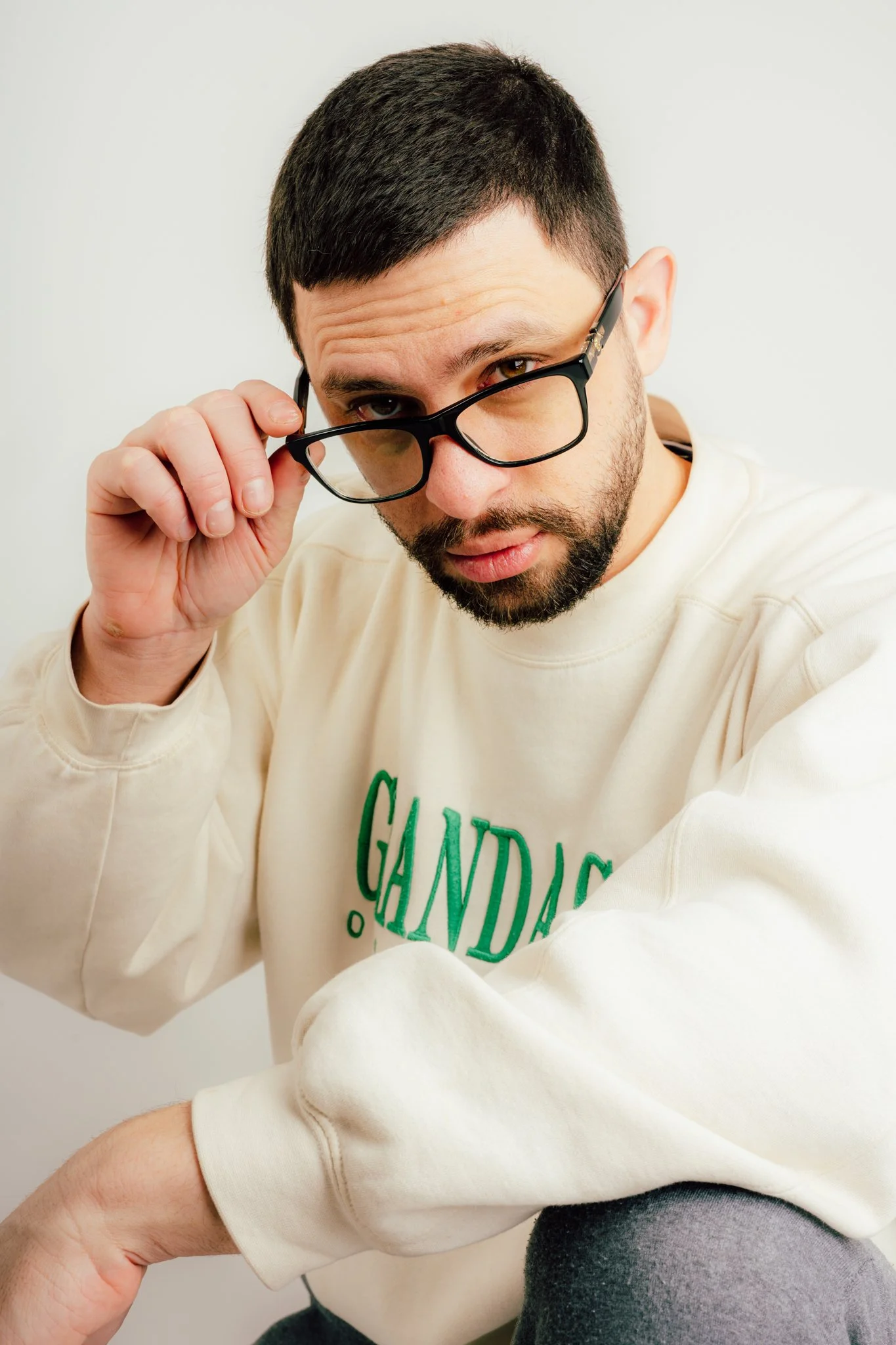 A young man with dark hair and a beard, wearing glasses and a cream-colored sweatshirt with green text, looks directly at the camera while adjusting his glasses in front of a plain white background.