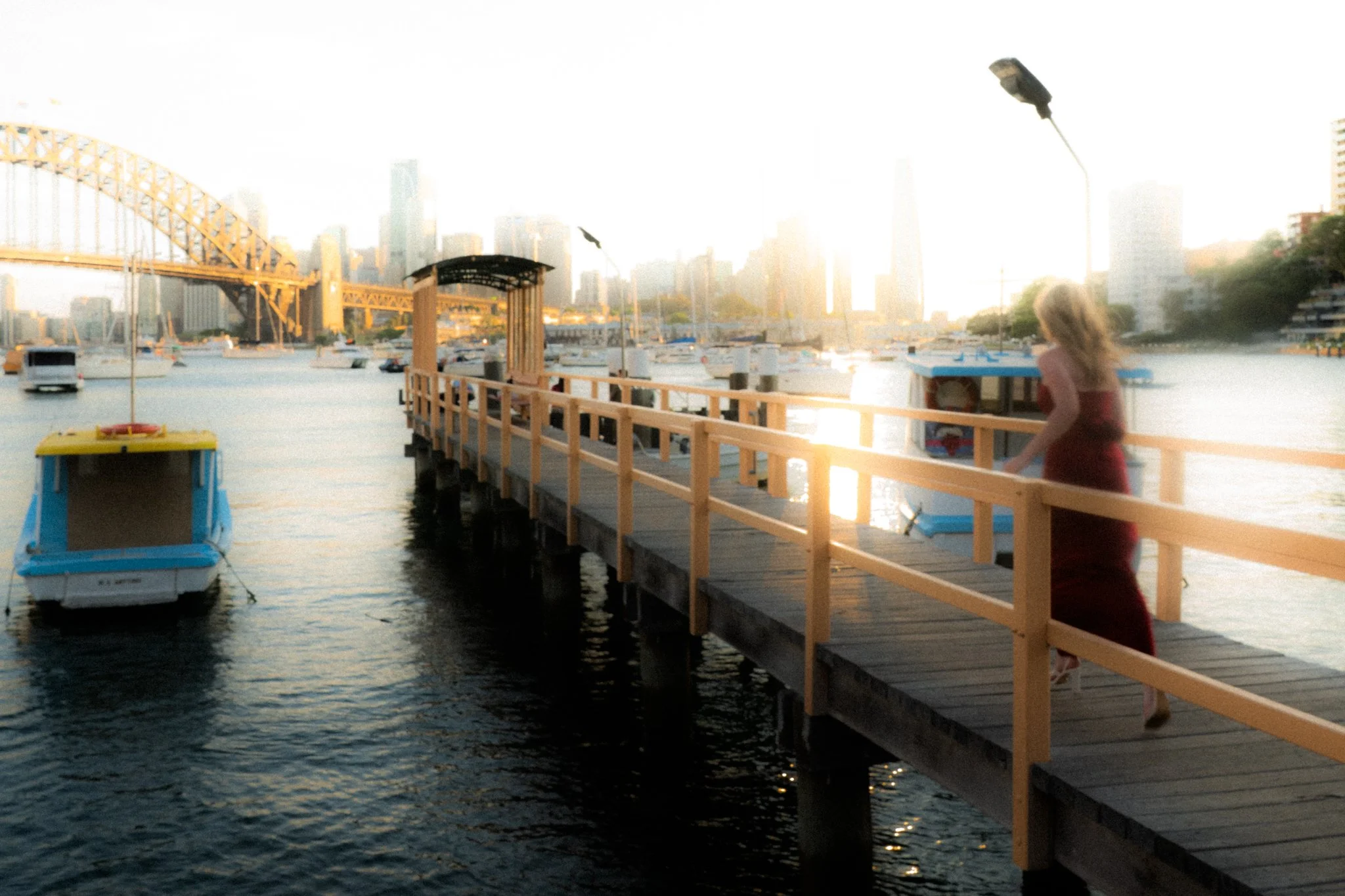 A woman in a red dress walking on a wooden dock by the water at sunset, with boats and city skyscrapers in the background.