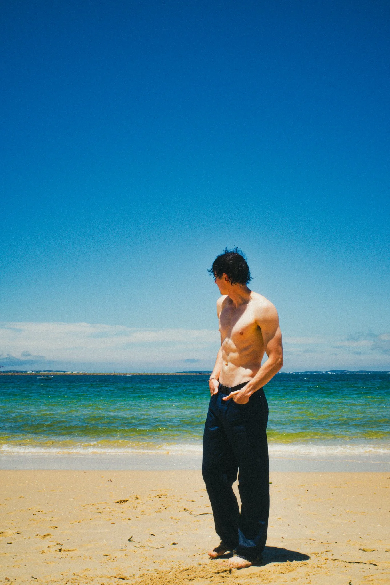 A shirtless young man with curly black hair standing on a sandy beach with the ocean and a clear blue sky in the background.