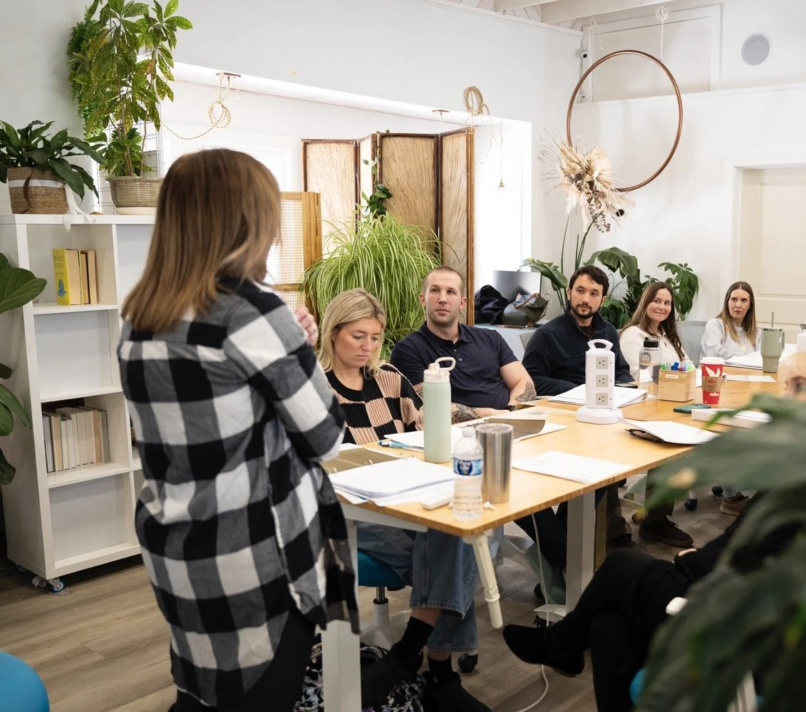 A woman in a checkered shirt is speaking to a group of five people seated at a table during a meeting in a bright, plant-filled room.