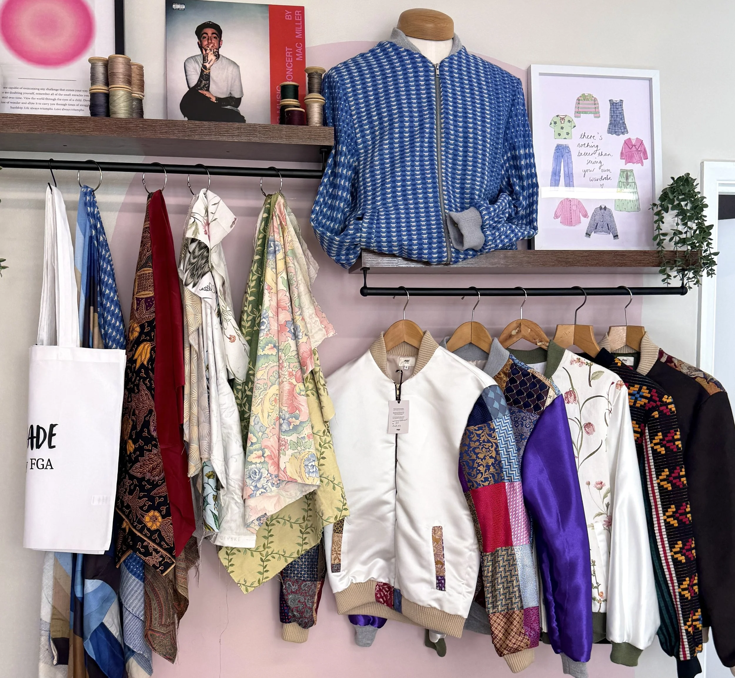 Display of a variety of patterned and solid-colored clothing items hanging on a rack, including jackets, shirts, and skirts, with a photo of a young man on a poster and sewing spools on the shelf above.