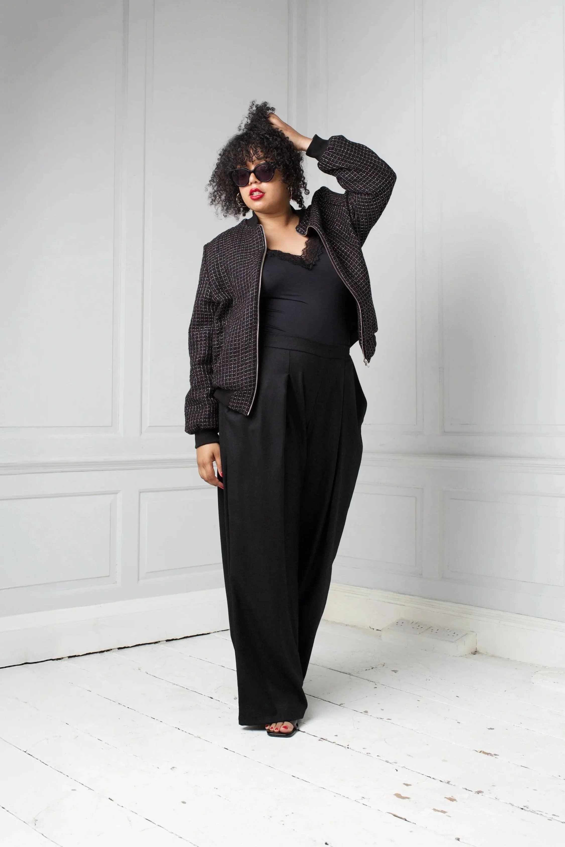 Woman with curly hair wearing sunglasses, a black lace-trimmed top, black pants, and a patterned jacket, standing in a white room with wood flooring.