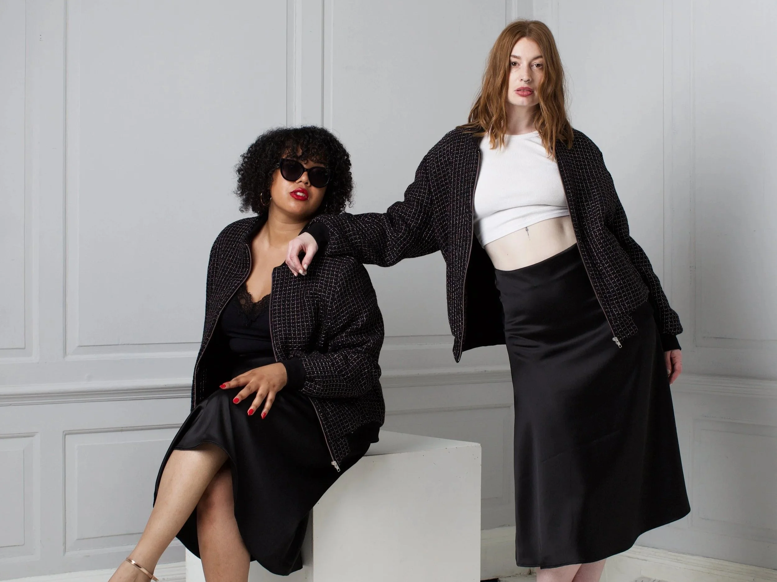 Two women in black and white outfits posing against a white wall. One woman is sitting with her leg crossed, wearing sunglasses and a black dress with a checked blazer. The other woman is standing, wearing a cropped white top and black skirt with a checkered jacket, her arm resting on the seated woman's shoulder.