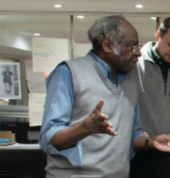 Two men are engaged in conversation in an office setting, one older with gray hair and glasses, wearing a gray vest over a blue shirt, and the other younger with dark hair, wearing a gray hoodie. They are standing near a desk with papers.