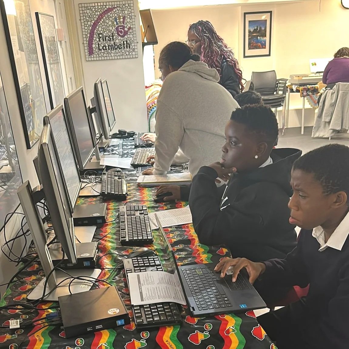 Several people working on desktop computers in a computer lab, with a woman standing and two young men focused on their screens.
