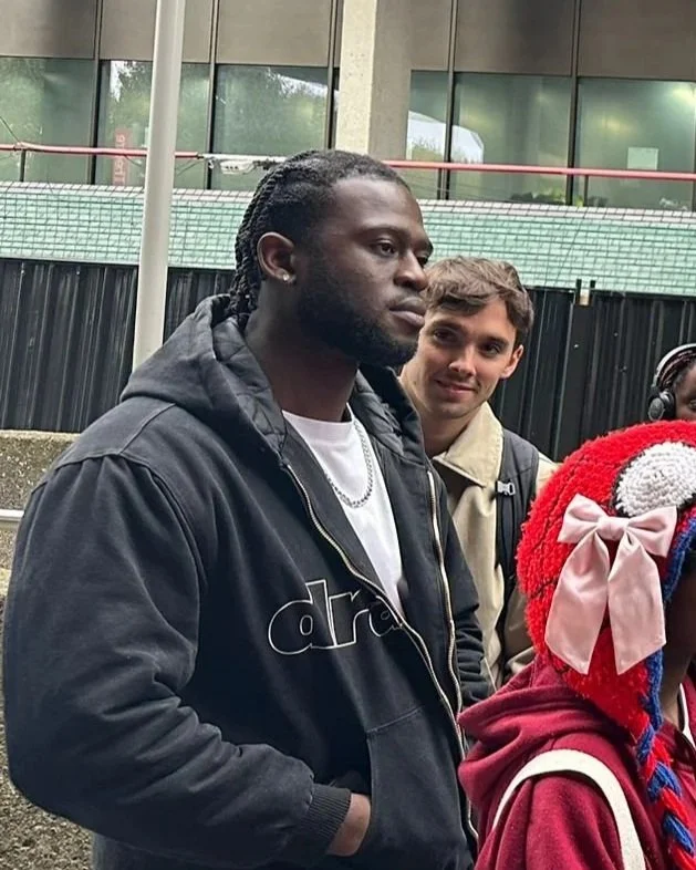 A group of people standing outdoors near a building, including a man with braided hair wearing a black hoodie and chain, a smiling man with light skin and brown hair wearing a beige jacket with a backpack, and a woman with a red hat with pink and white bows, and a red hoodie.