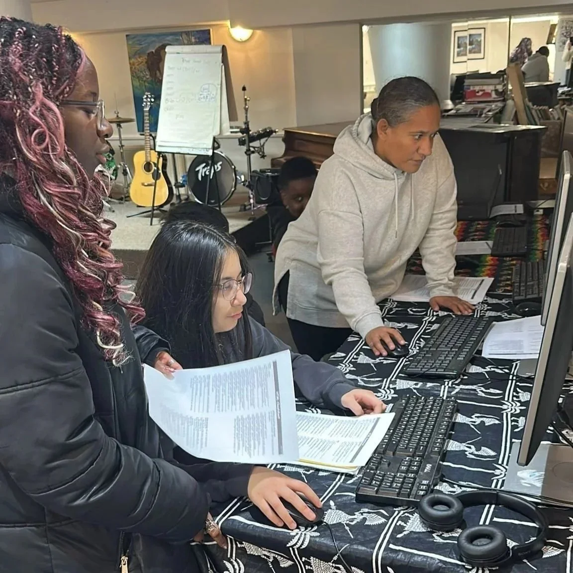 Three women working at computer stations, one woman with glasses and long dark hair looking at a document, another woman in freestyle hair and glasses focused on her screen, and a woman with a grey hoodie standing and working on a computer. Musical instruments such as a guitar and drum set are in the background.