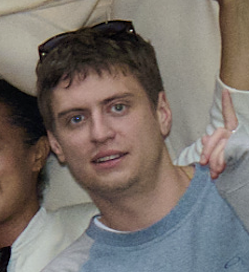 Close-up of a young man with short brown hair, wearing a gray T-shirt, and sunglasses on his head, making a peace sign with his hand.