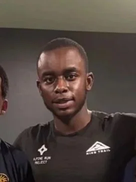 A young man with dark skin and short hair, wearing a black athletic shirt with white logos, standing indoors against a dark wall.