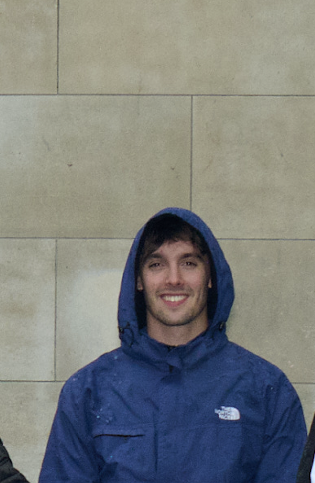 Young man in a blue North Face jacket with a hood, smiling, standing against a beige stone wall.