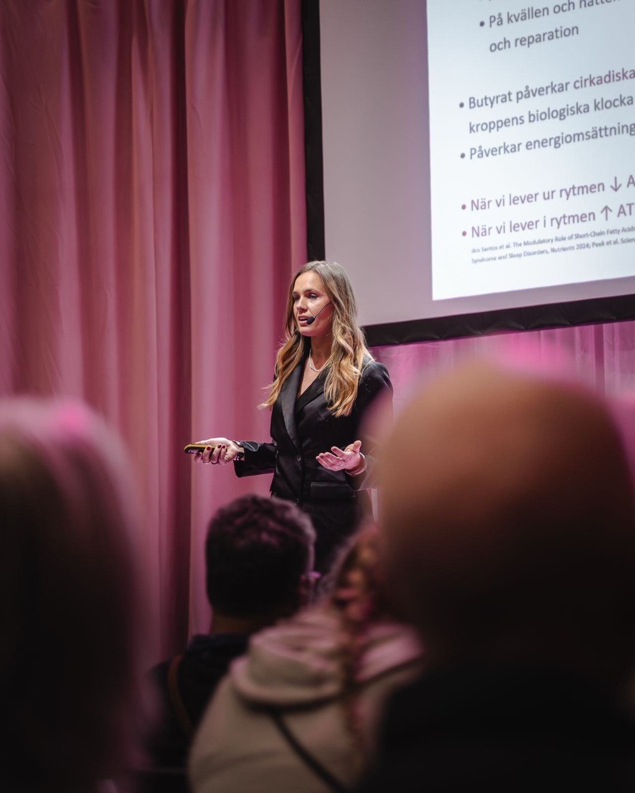 Kvinna som håller en presentation framför en skärm med text, i en salong med rosa gardiner och en publik.