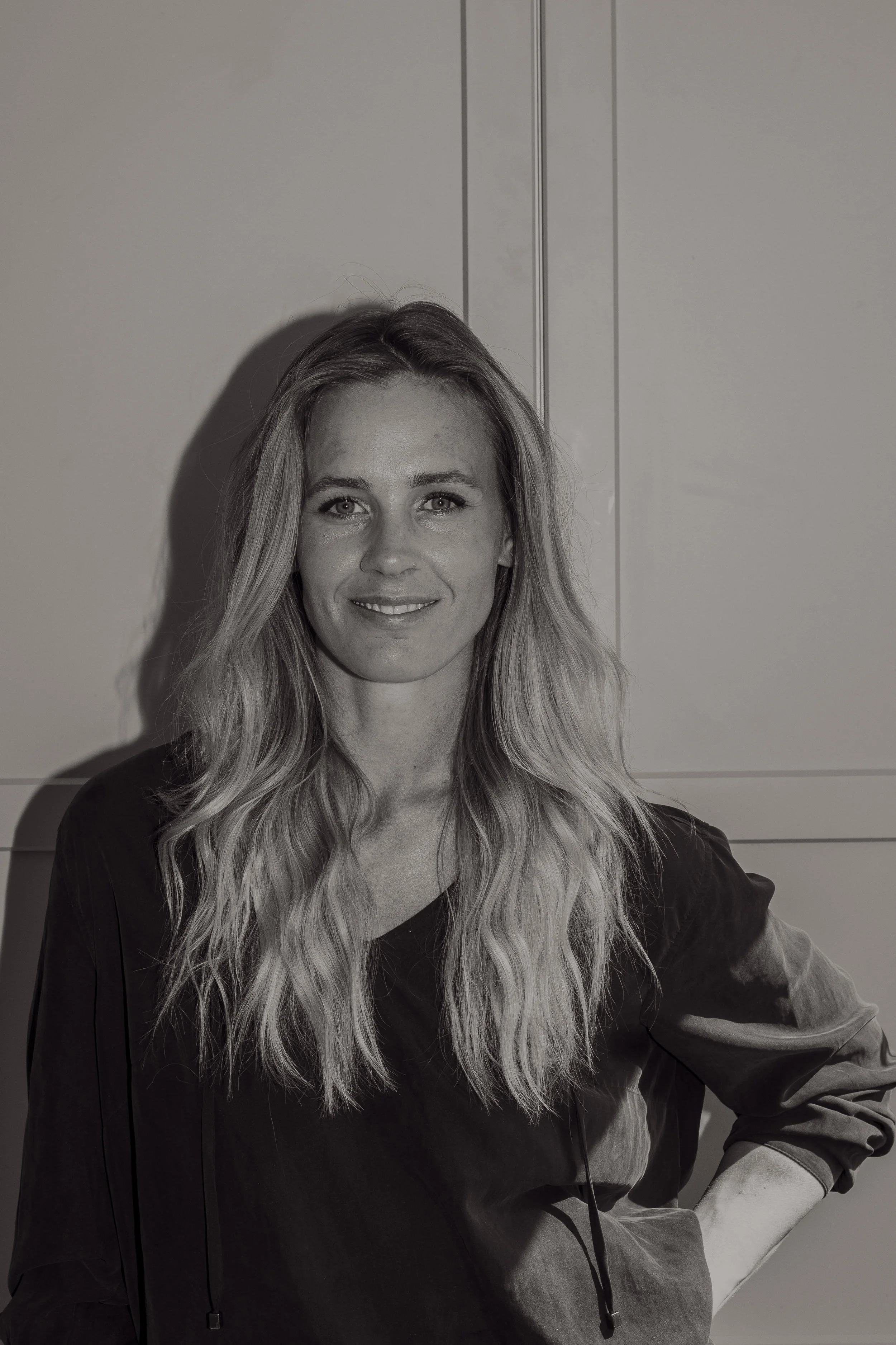 Kvinna med långt, blont, lockigt hår ler mot kameran, står mot en vägg.