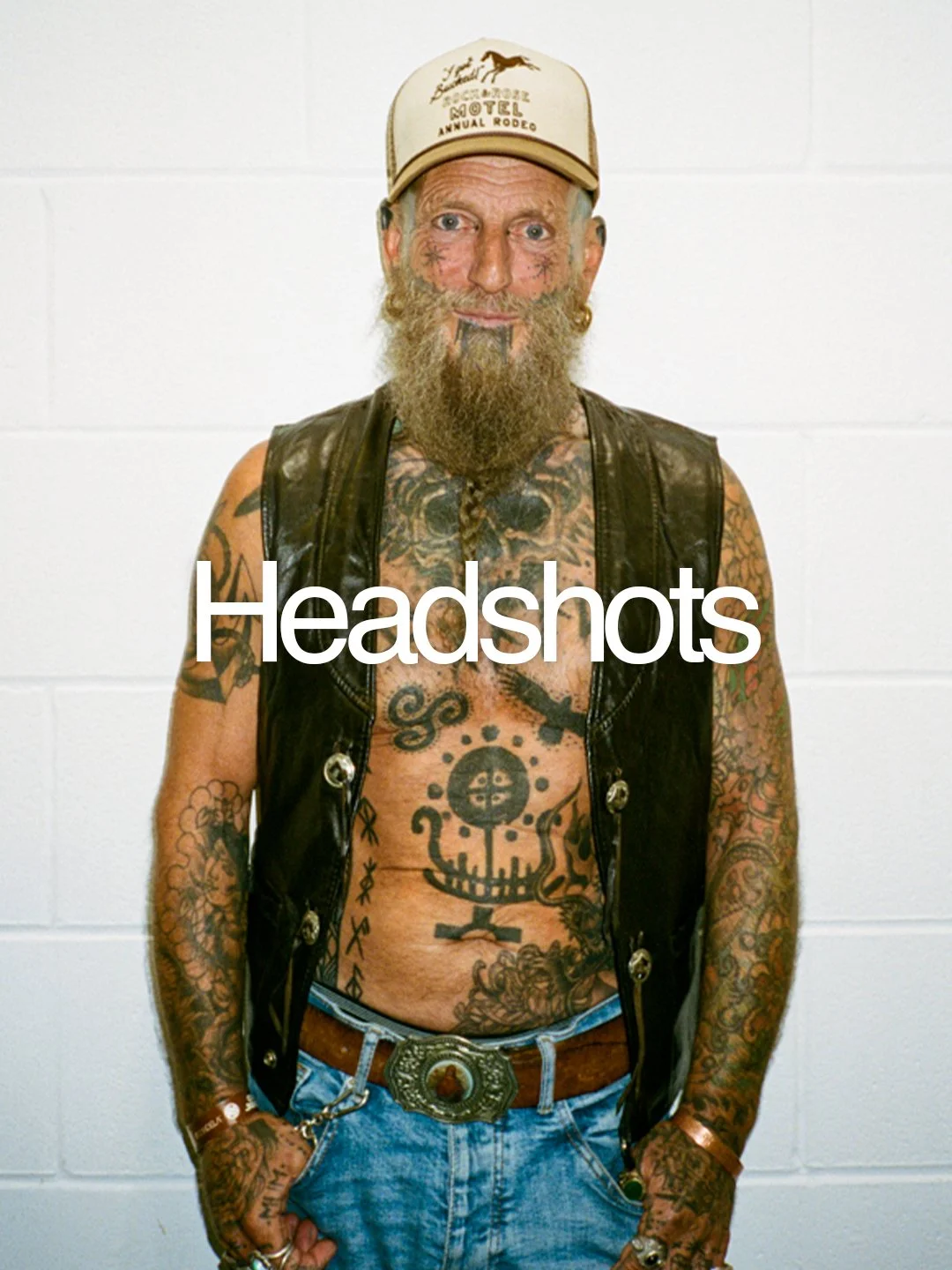 A tattooed man with a beard, wearing a beige trucker hat, black leather vest, and denim jeans with a large belt buckle, standing against a white wall.