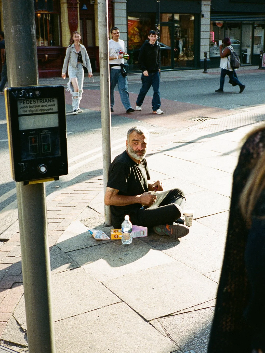 A man sits on the sidewalk at a pedestrian crossing with various items around him, including a bottle of water, a box, and a cup. In the background, pedestrians walk across the street, and there is a traffic signal showing red and green lights for pe
