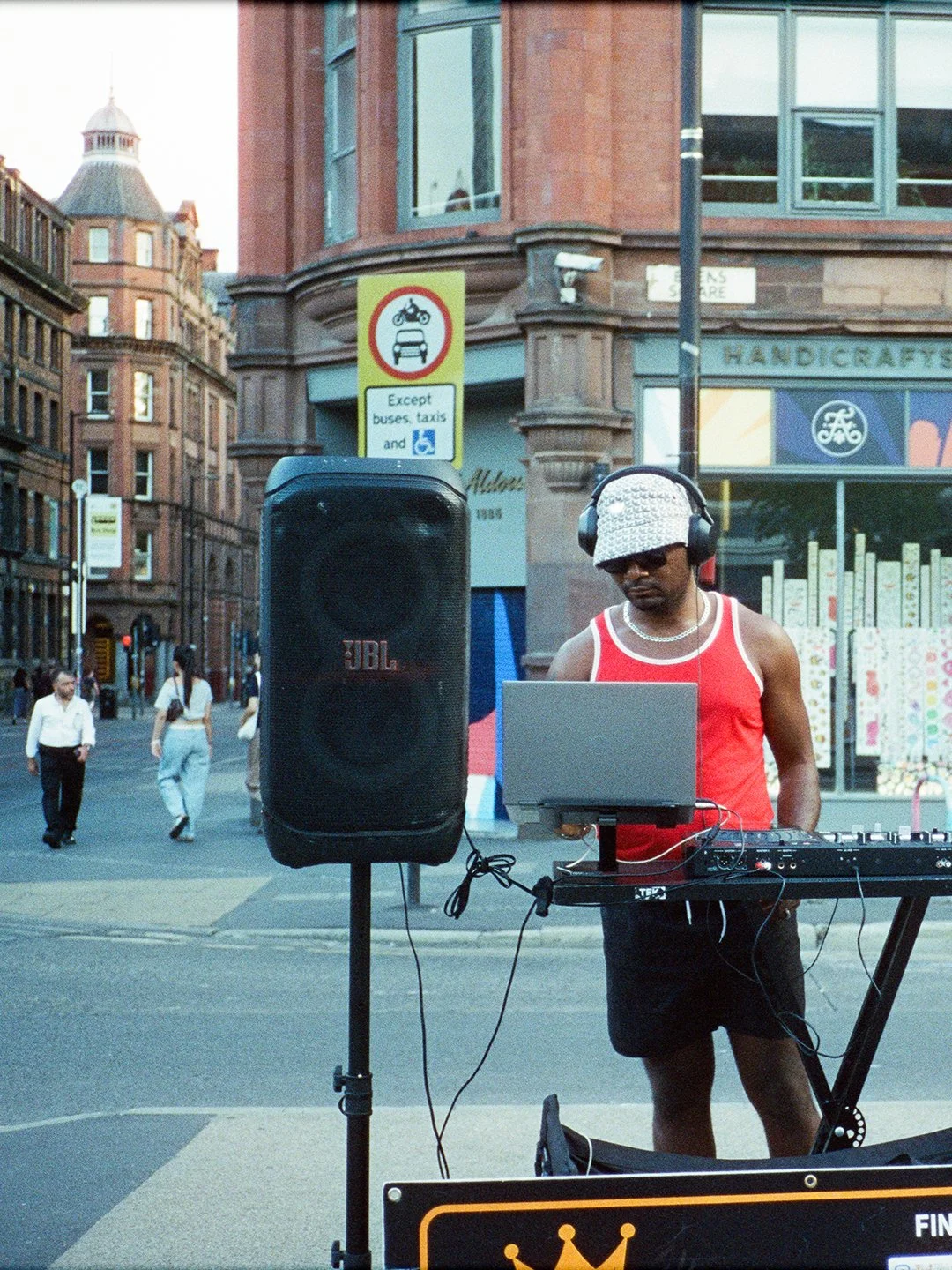 A DJ wearing a white patterned hat, sunglasses, and a red tank top performing at an outdoor city street event, with a speaker, laptop, and mixing equipment in front of him.