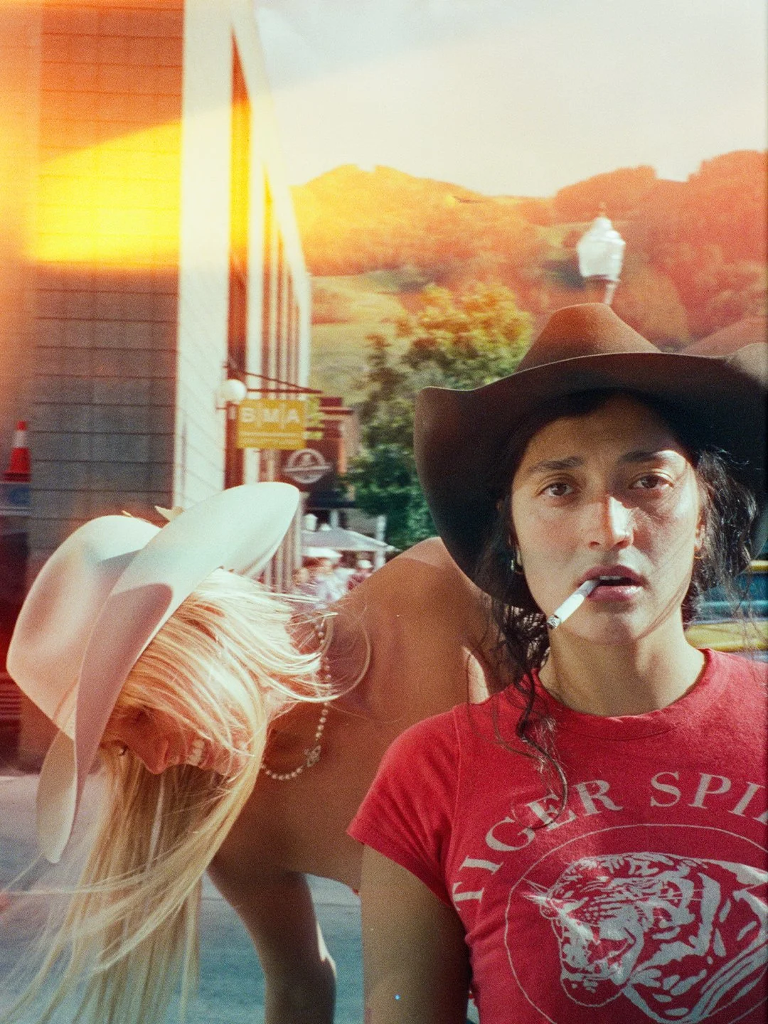 A woman wearing a cowboy hat and red Tiger Spirit t-shirt, with a cigarette in her mouth, standing in front of a blonde woman with a large white hat, in an urban outdoor setting with a hilly background.