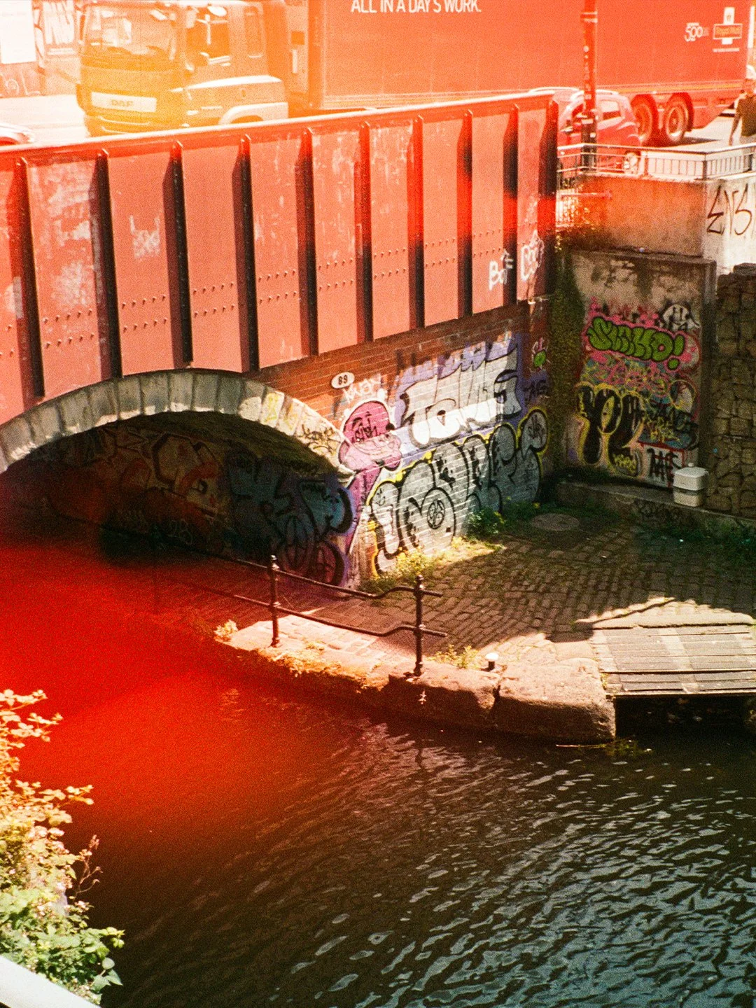 A canal under a brick bridge with graffiti on the walls, and a cobblestone path beside the water. A red truck and two cars are visible on a nearby street.